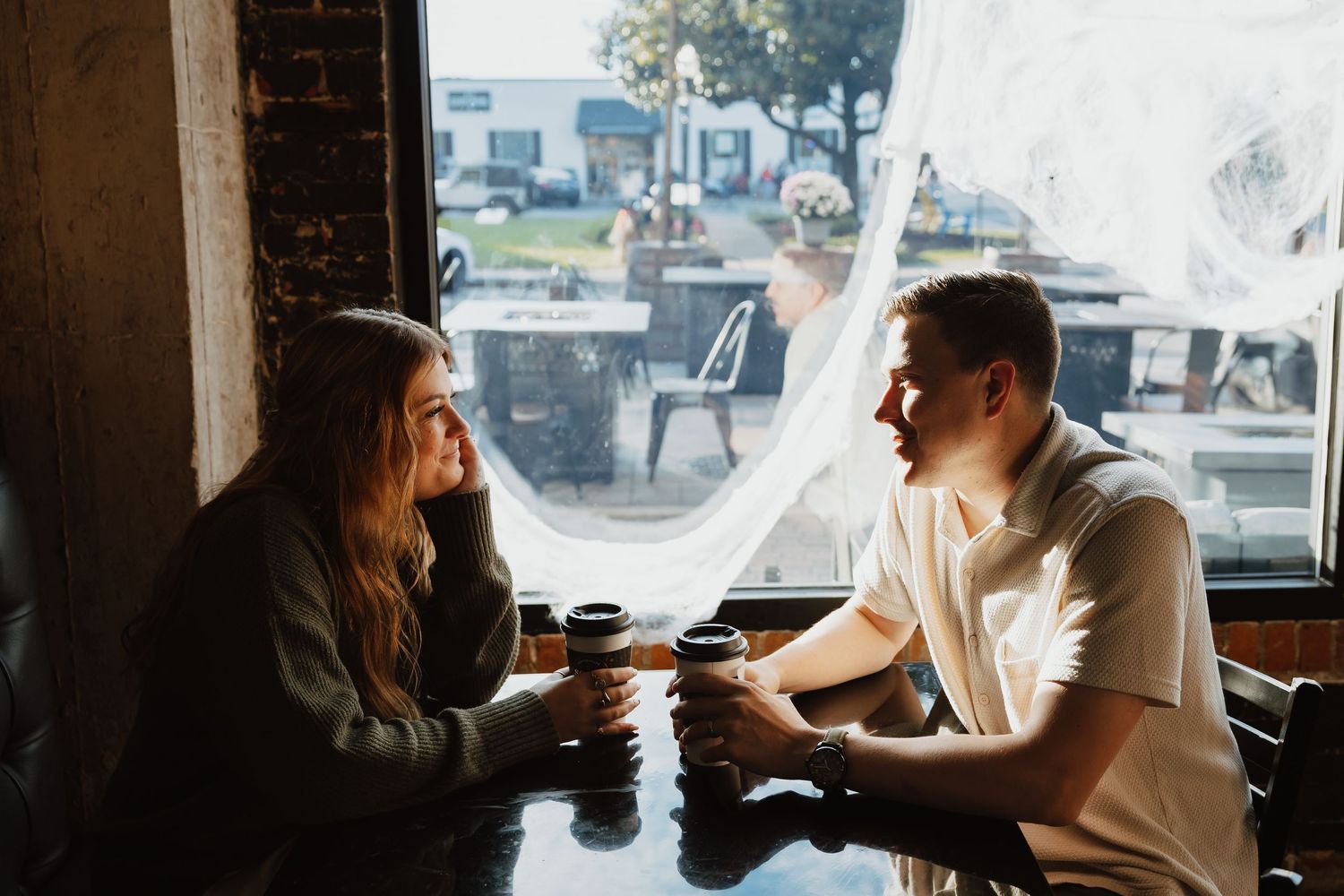 Two people having coffee and conversation at a cafe table by a window in warm sunlight.