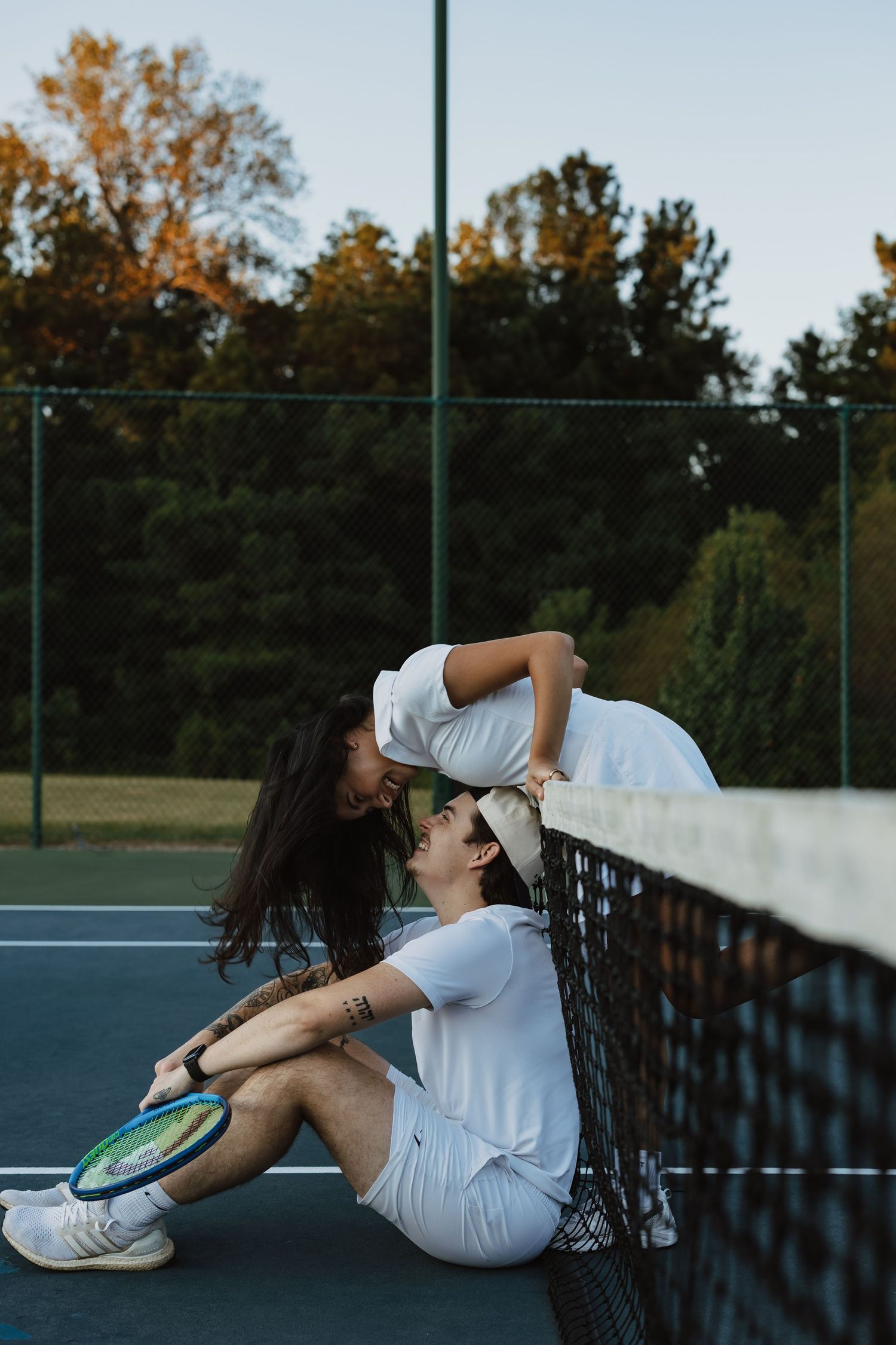 People in tennis attire relax and pose along a tennis court net during golden hour.