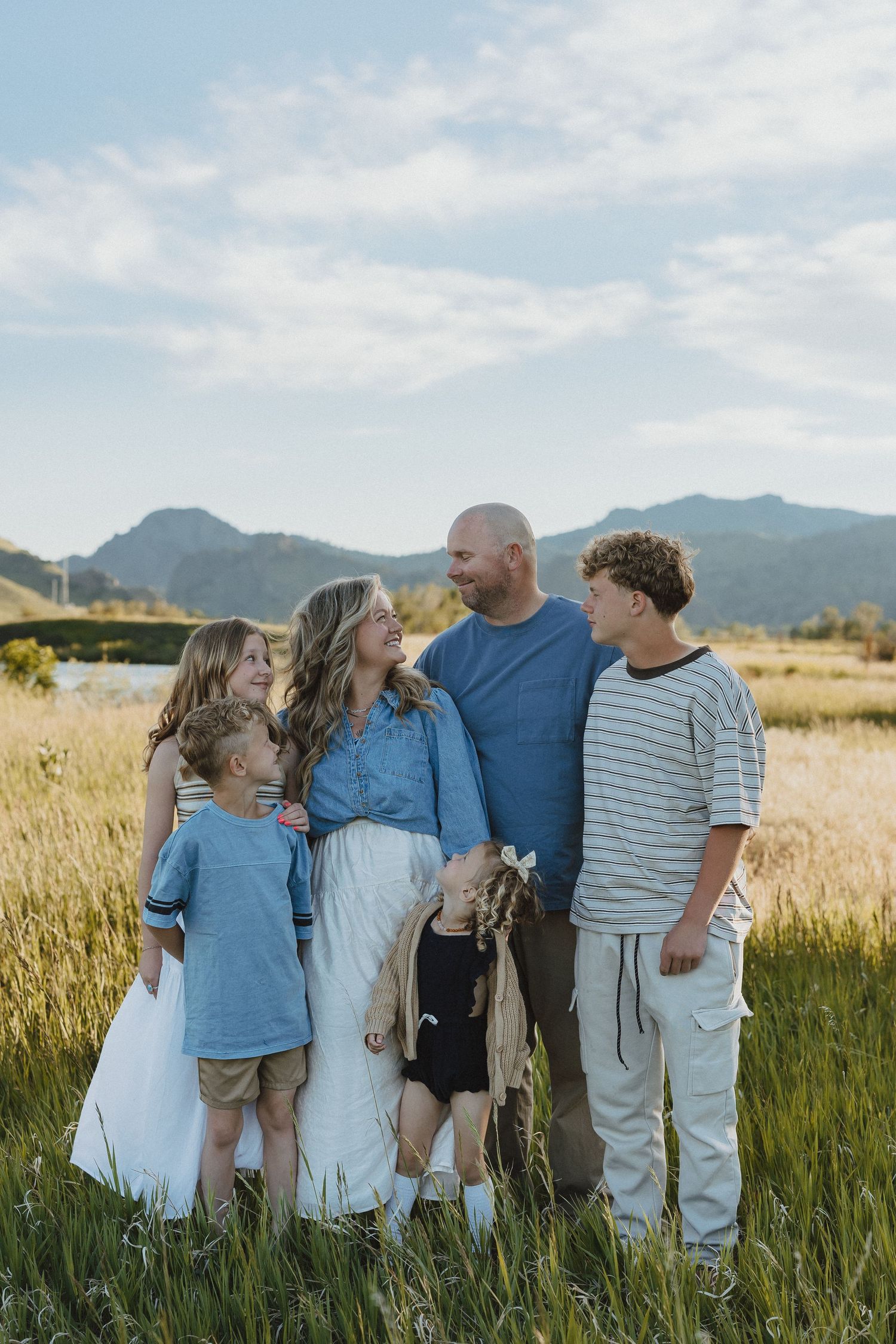 Family of five smiling together in a golden grass field with mountain backdrop on a sunny day.