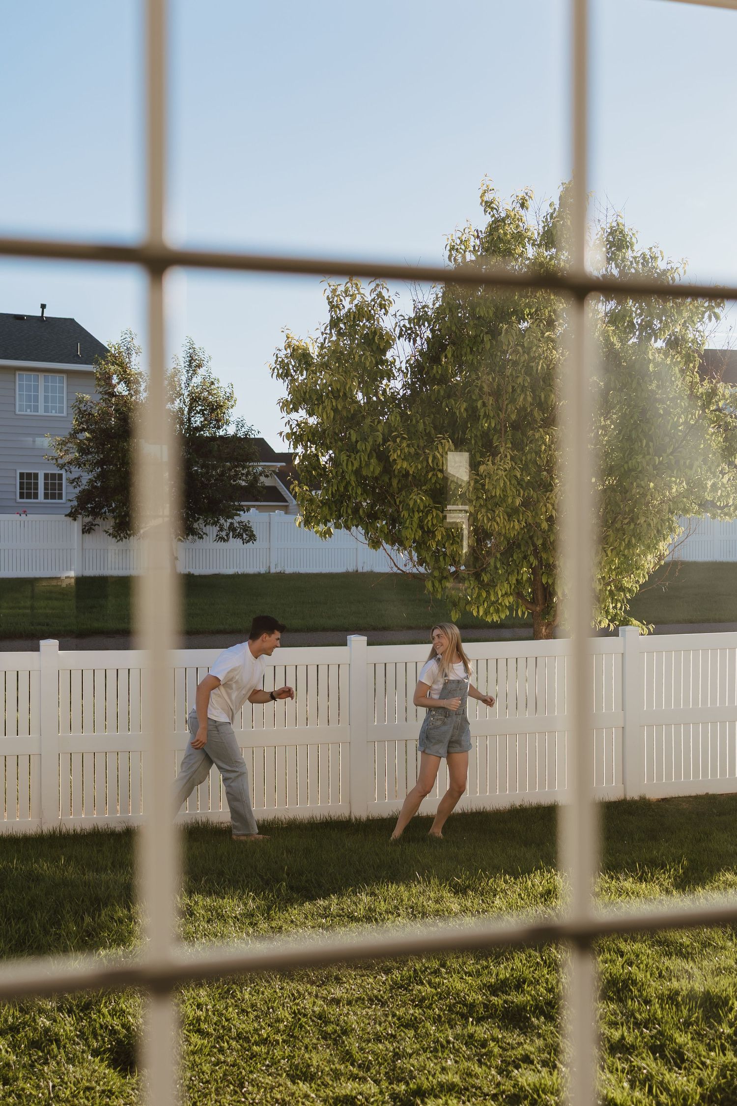 Two children playing in a sunny backyard with a white picket fence, viewed through a window screen.