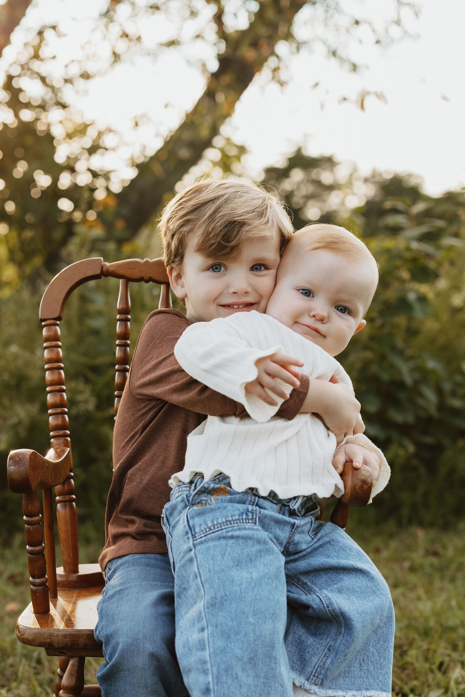 Children sitting together on a wooden chair outdoors with greenery and sunlight in the background.