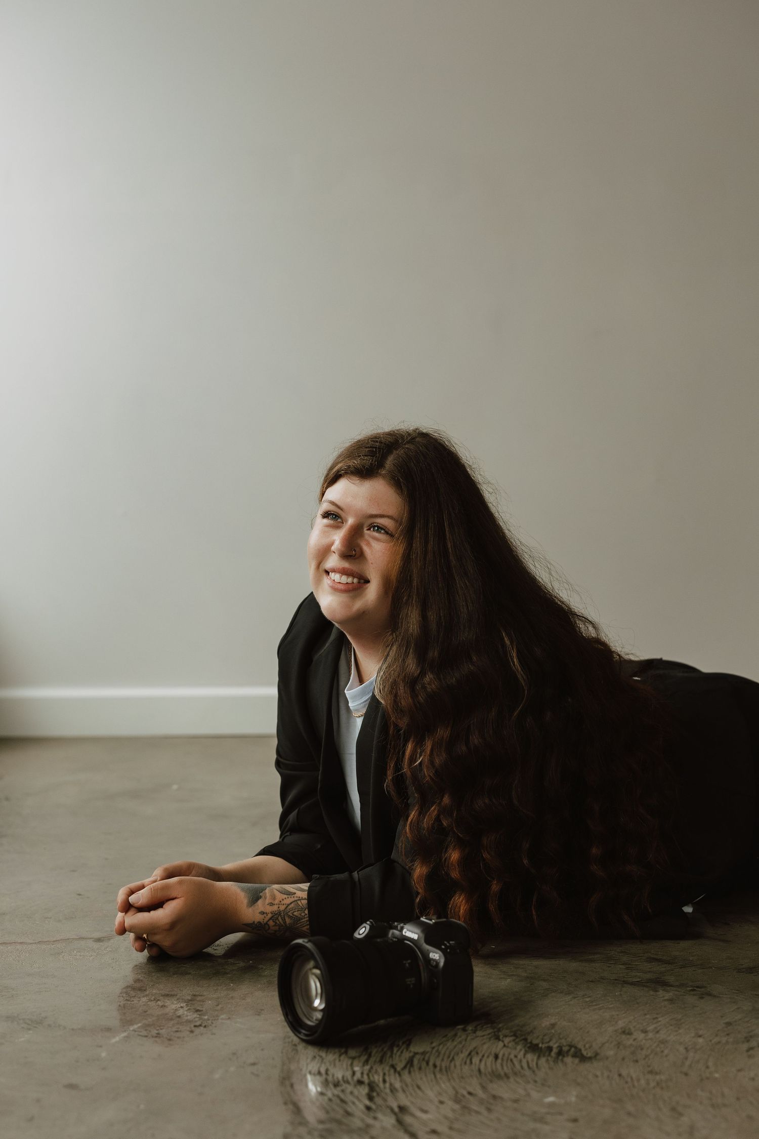 A photographer lies on concrete floor with a professional camera, smiling and wearing a black jacket.