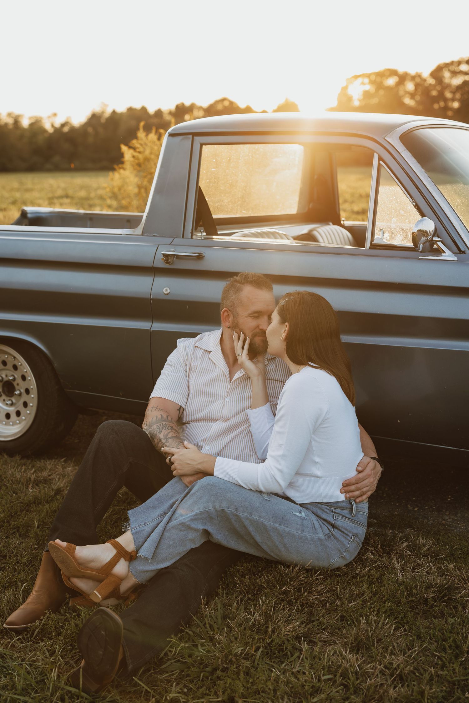 A couple relaxes together on grass beside a classic car during golden hour sunset.