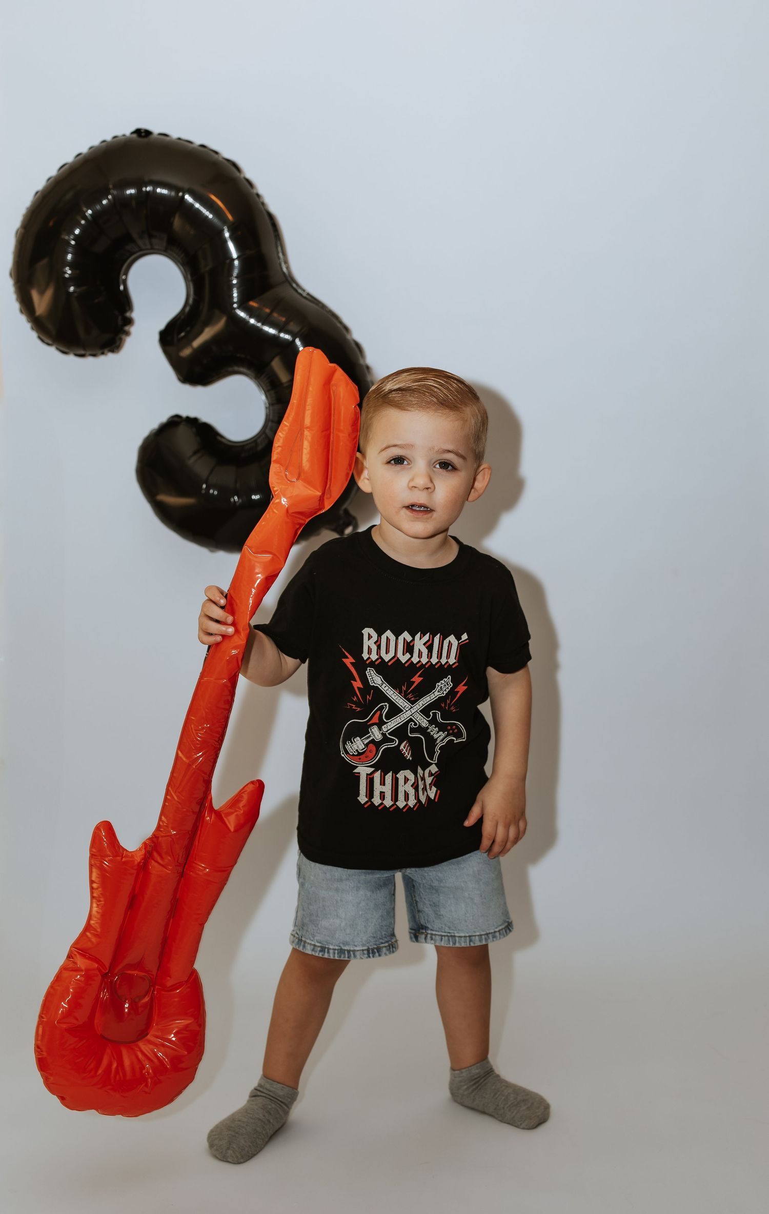 A toddler in a black rock and roll t-shirt and denim shorts poses with red inflatable guitar props against a white wall.