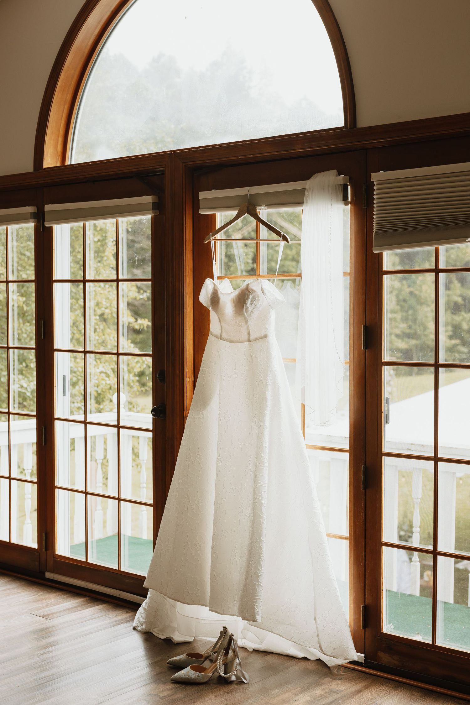 White wedding gown hanging on wooden hanger in front of arched French doors with heels below.