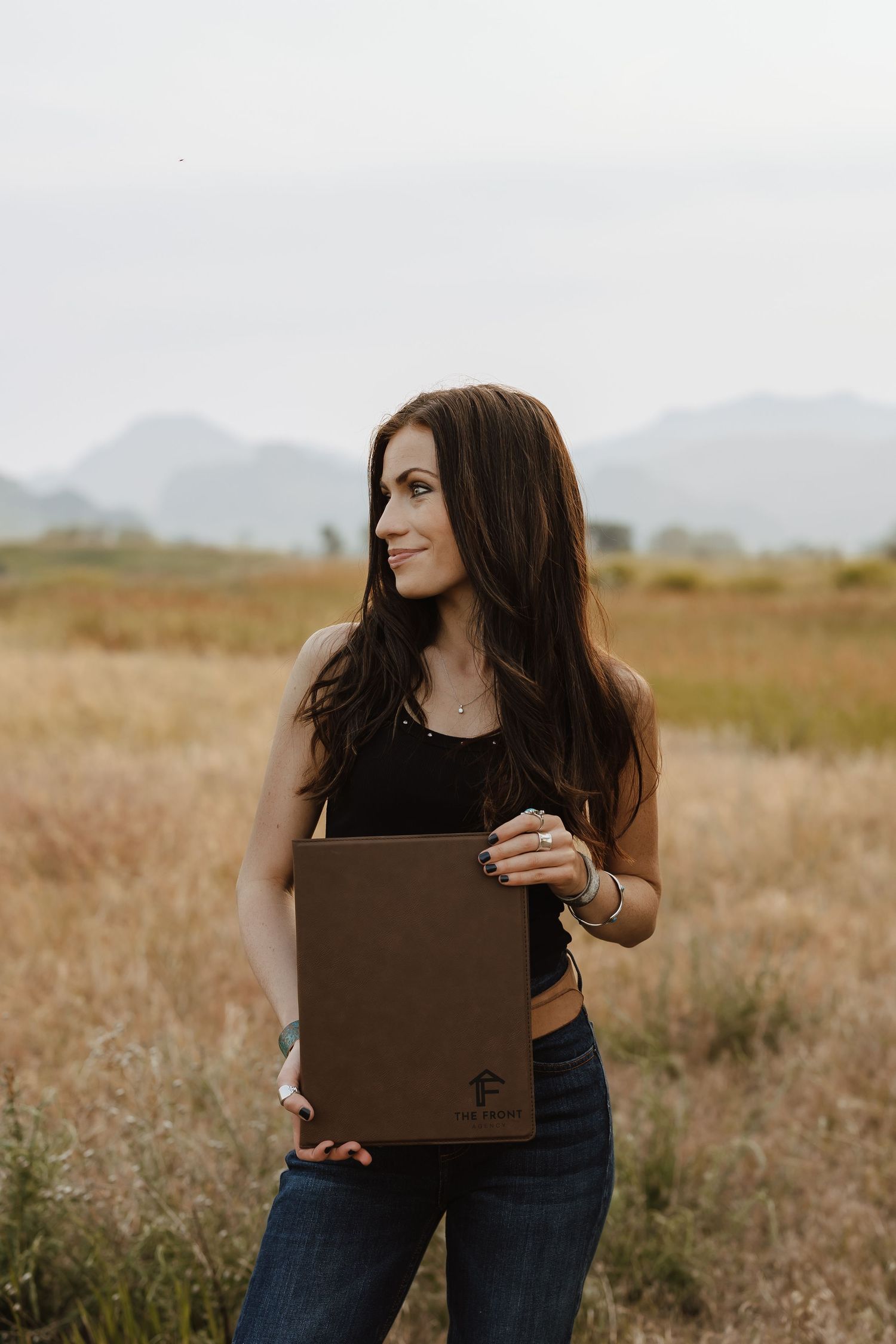 Woman with long dark hair holding a brown leather portfolio in a golden field with mountains in the background.