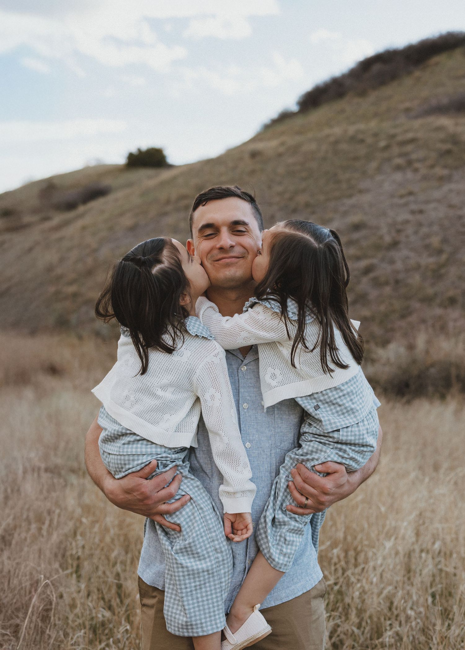 A father holds two young girls in a field, both daughters kissing his cheeks in a heartwarming outdoor portrait.