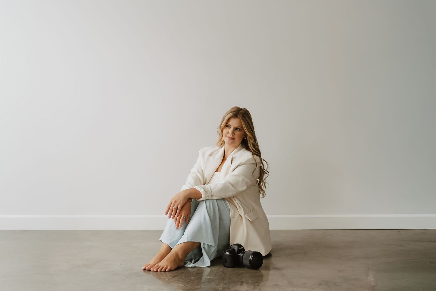 Woman in white dress sits gracefully on floor in minimalist studio setting with soft natural lighting.