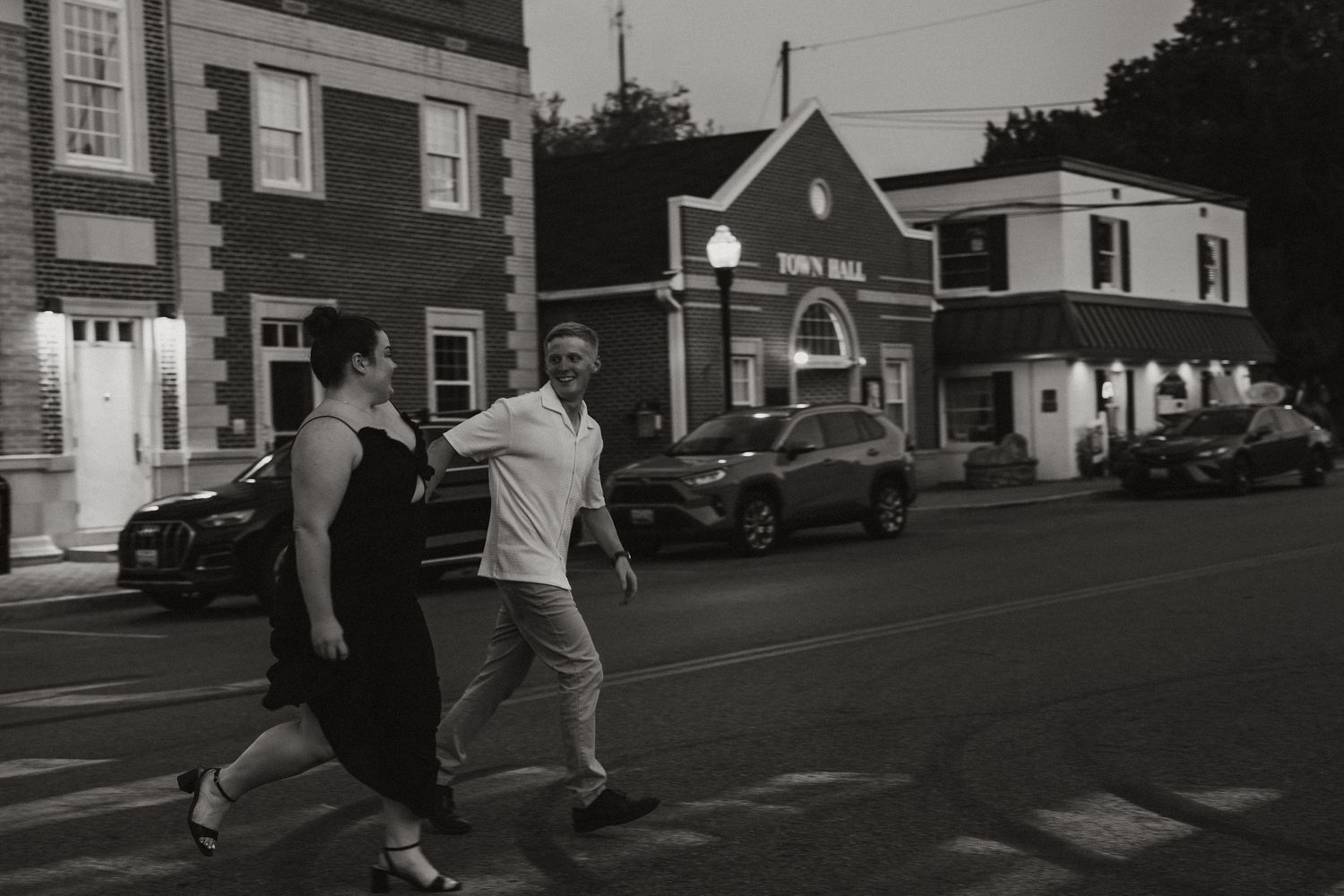 People cross a small town street at dusk with vintage cars parked along storefronts in black and white photography.