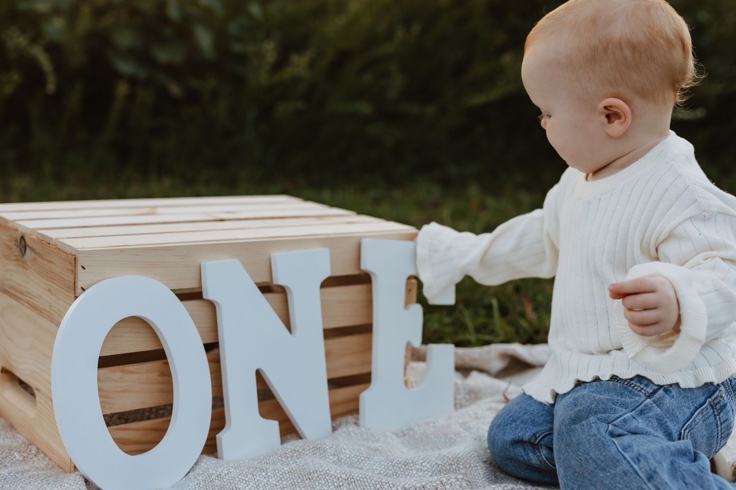 A baby in white clothing sits on wooden steps next to decorative white letters spelling out a word.