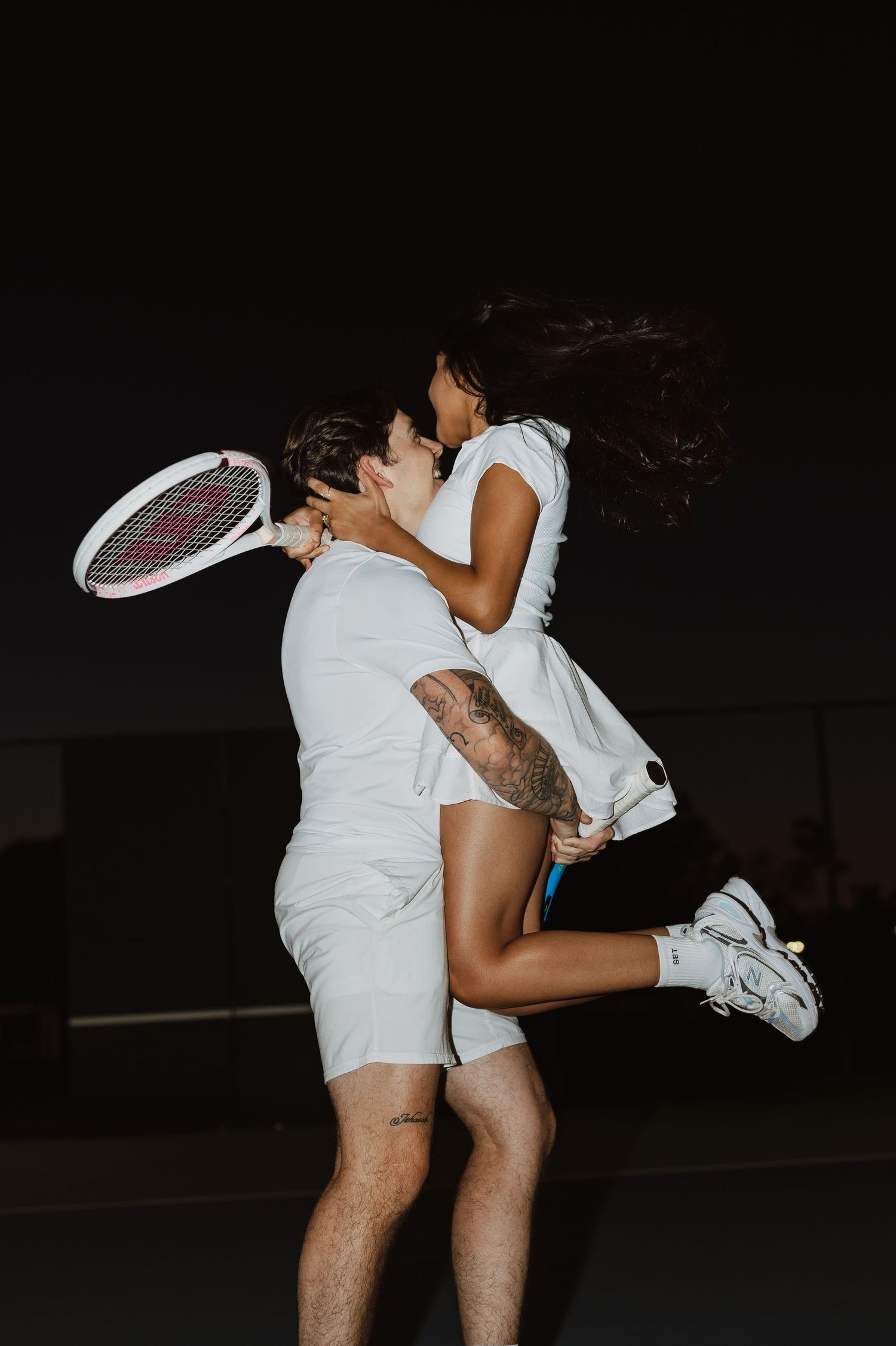 Two people playing tennis at night on a dark court wearing white outfits and using tennis rackets.