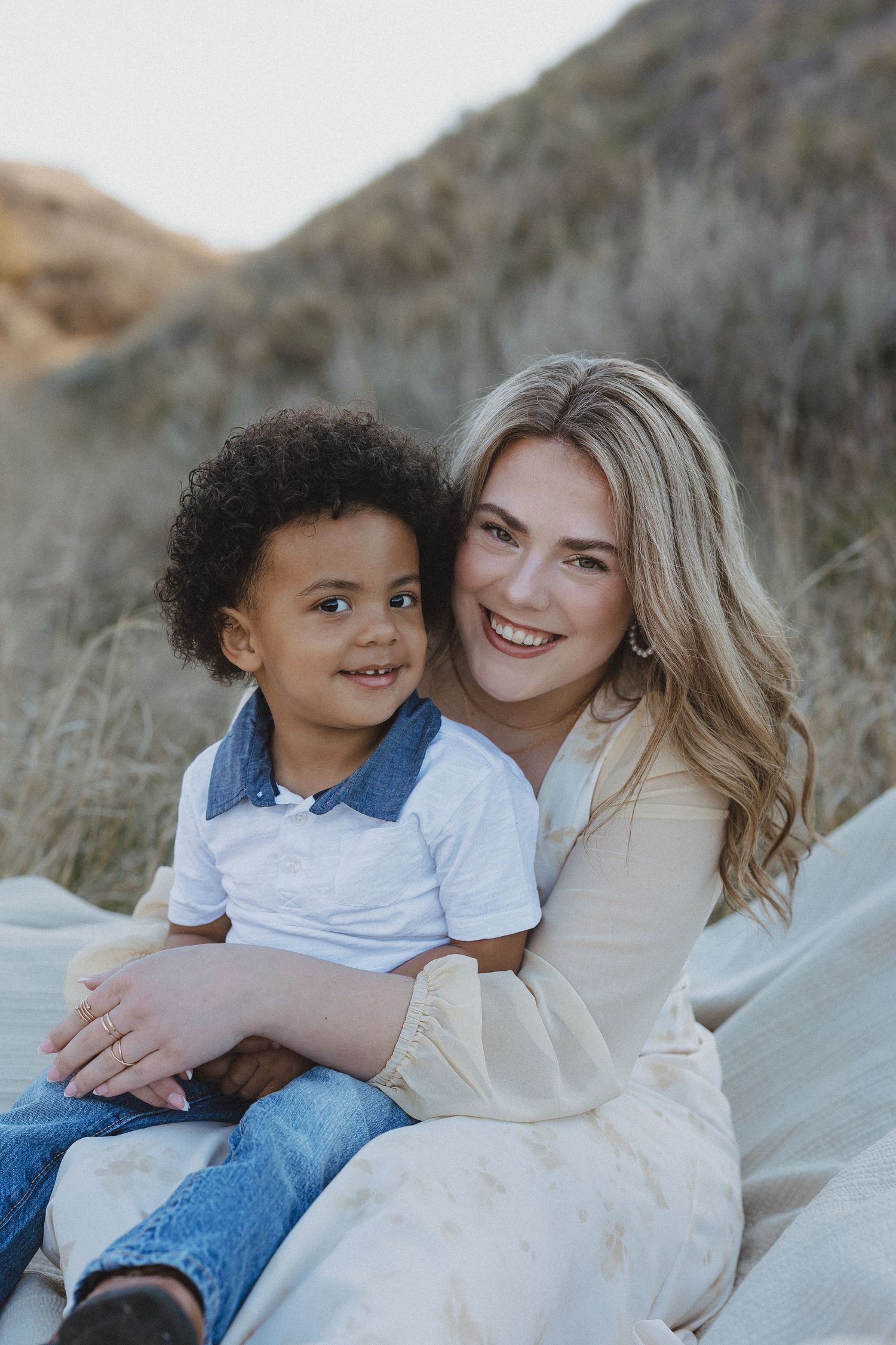 A smiling blonde woman hugs a young curly-haired boy outdoors on a blanket with grassy hills behind them.
