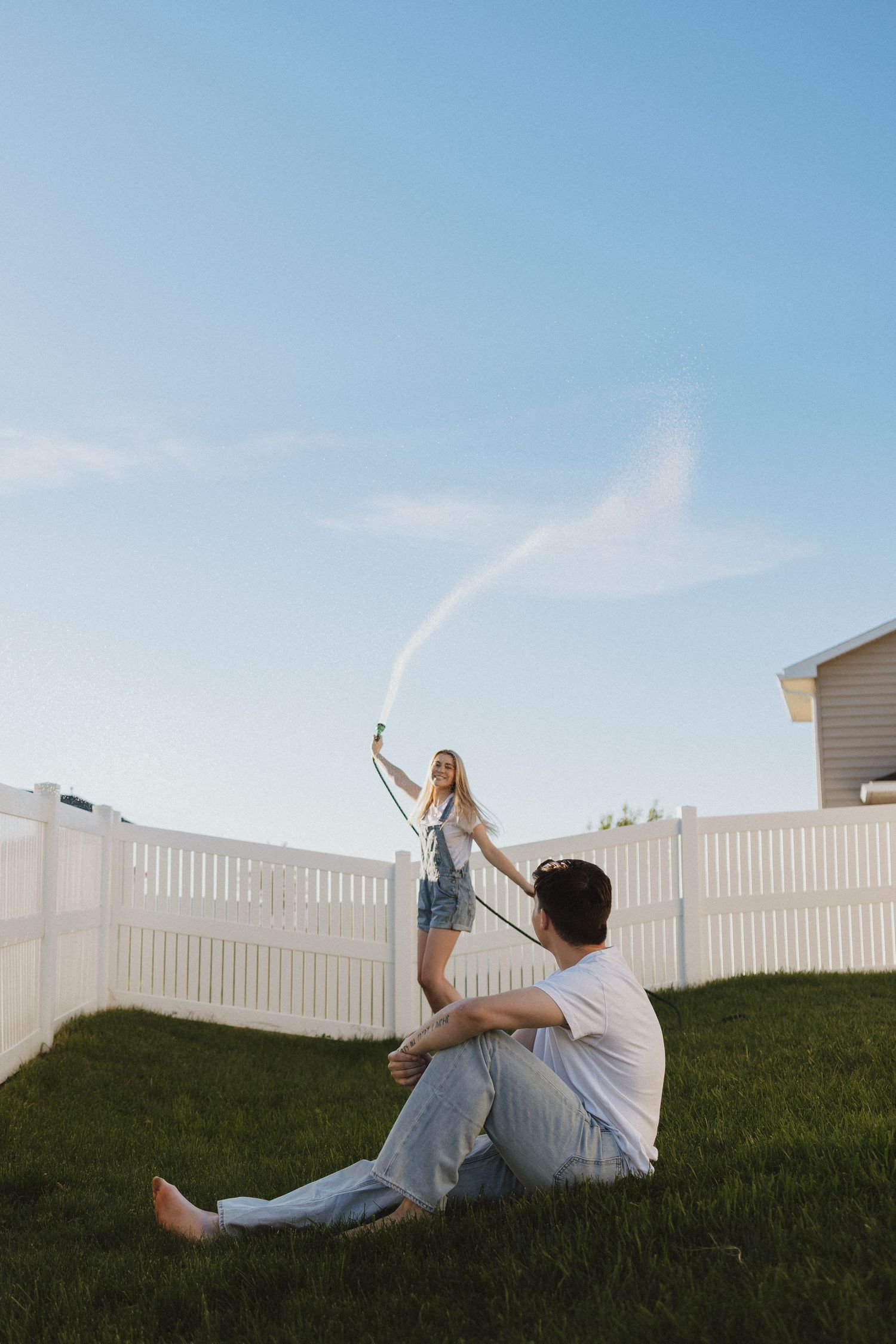Woman spraying water hose in backyard while man sits on grass near white vinyl fence on sunny day.