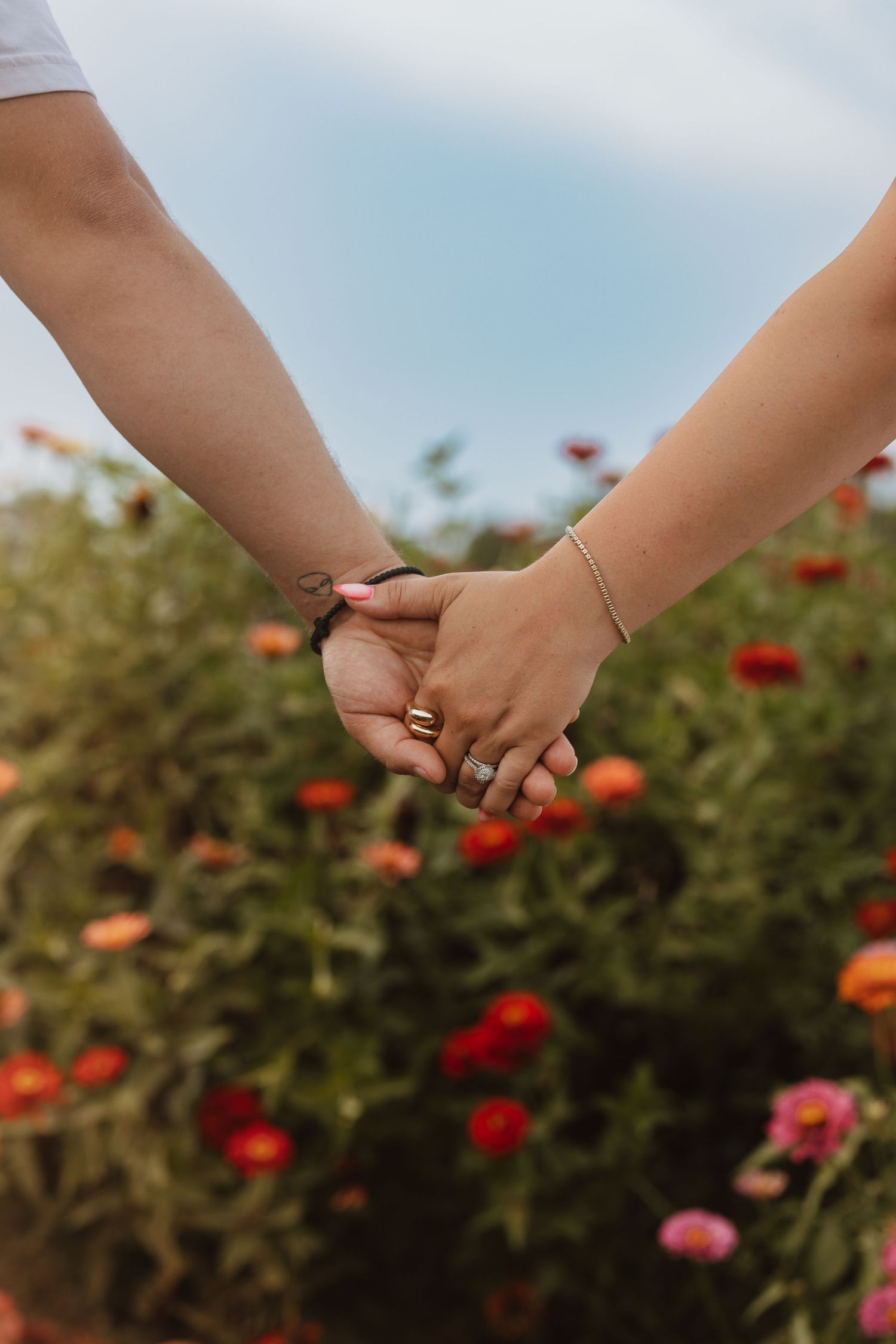 Two people holding hands in a garden with blooming red and pink flowers against a soft blue sky.