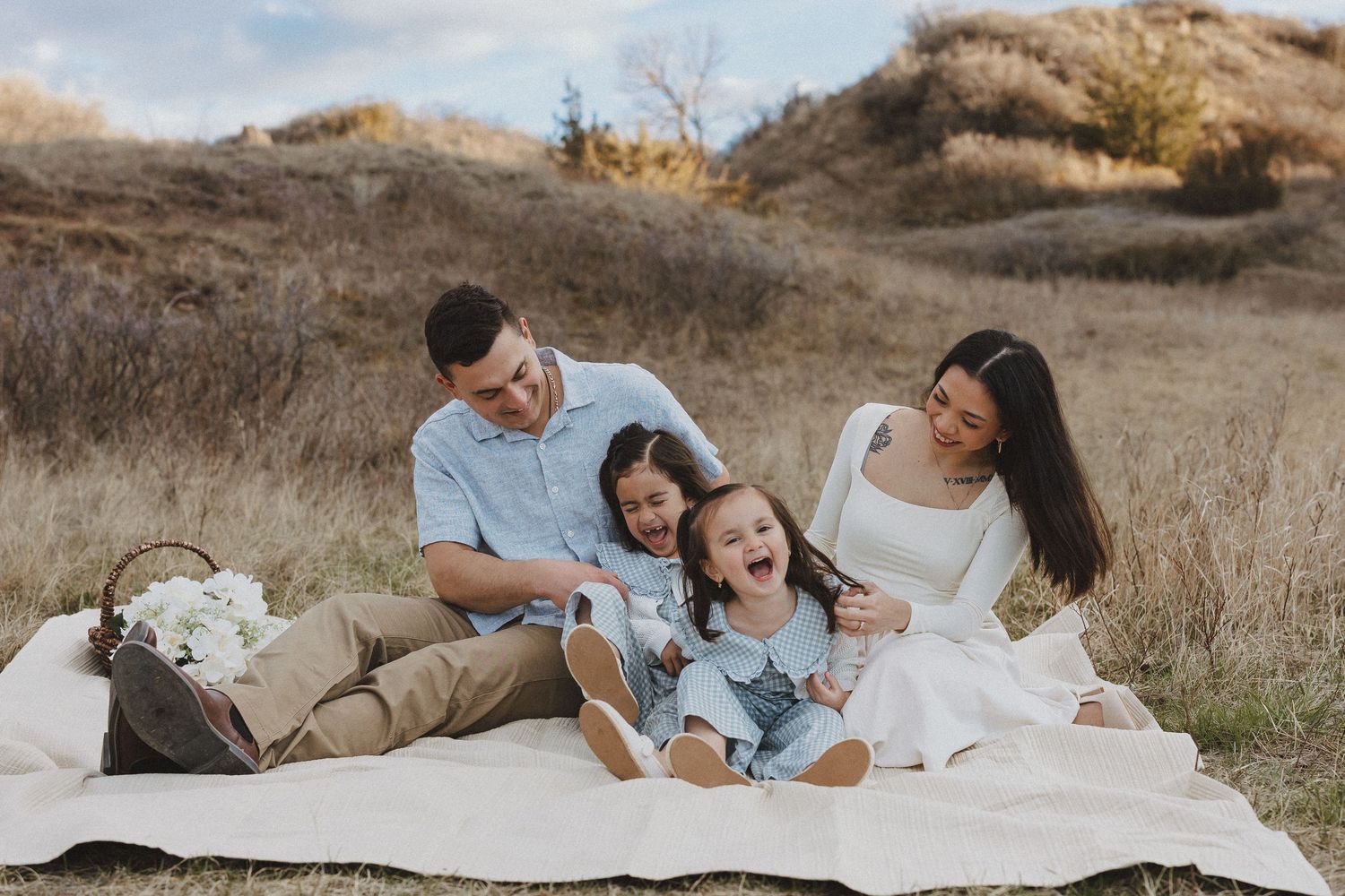 Happy family of four laughing together on a blanket in a golden field during an outdoor photo session.