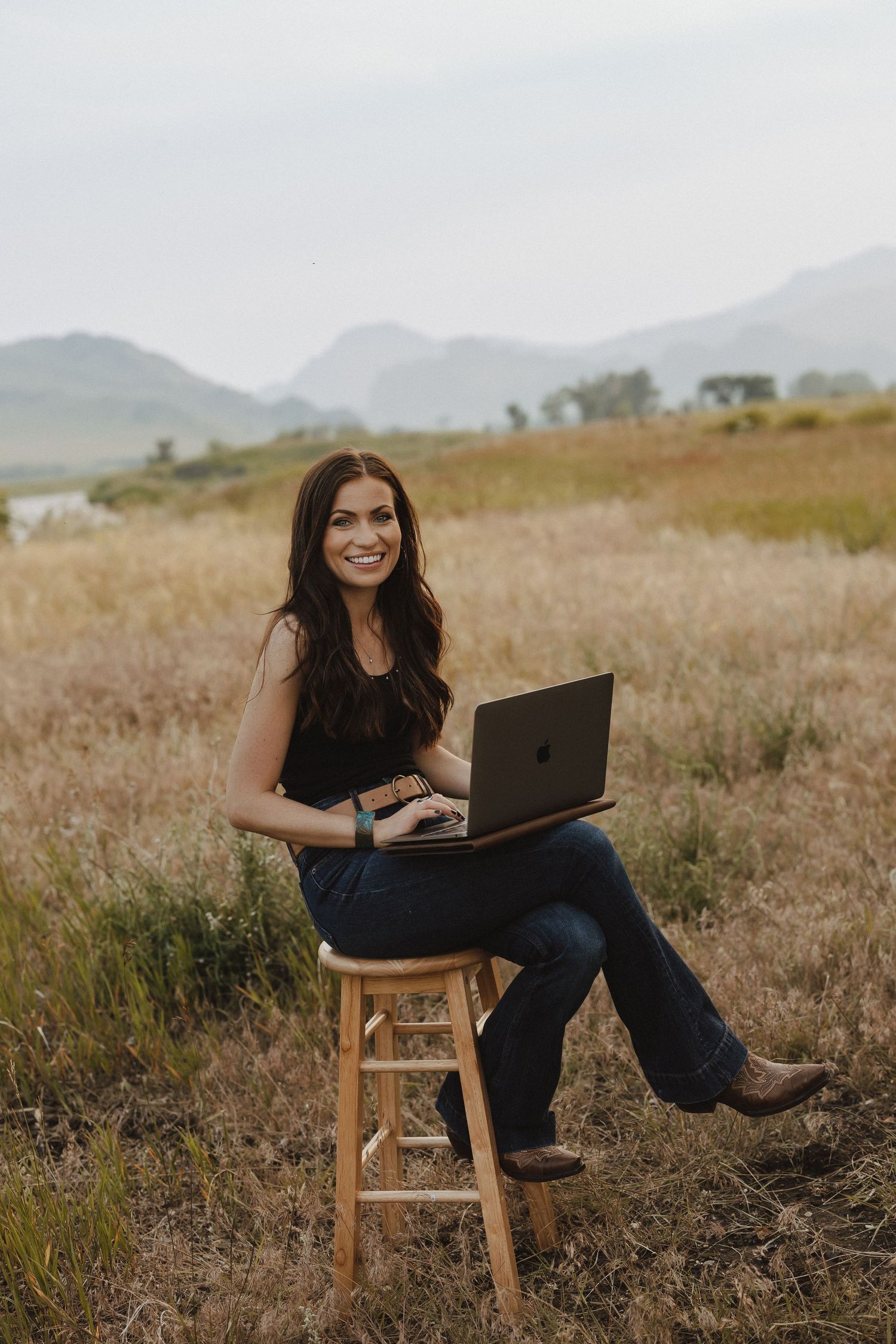 Smiling brunette woman sitting on wooden stool with laptop in scenic mountain field wearing black top and jeans.