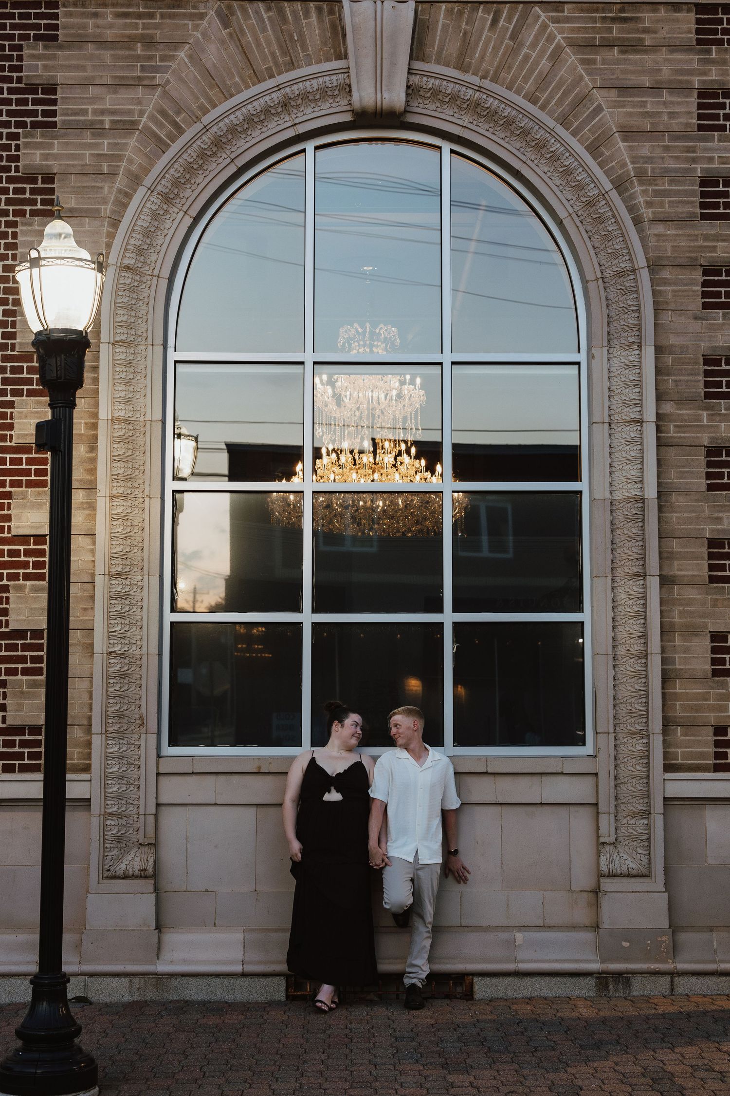 A couple stands in front of an arched brick window with a chandelier reflection and vintage lamp post at dusk.