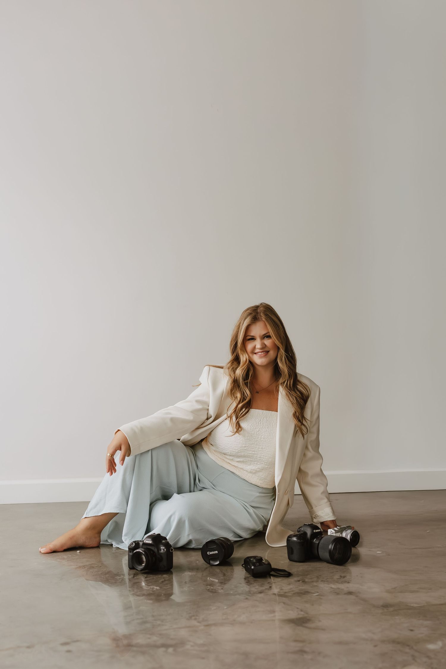 A person in a cream blazer and light dress lounges on a concrete floor in a minimalist studio setting.