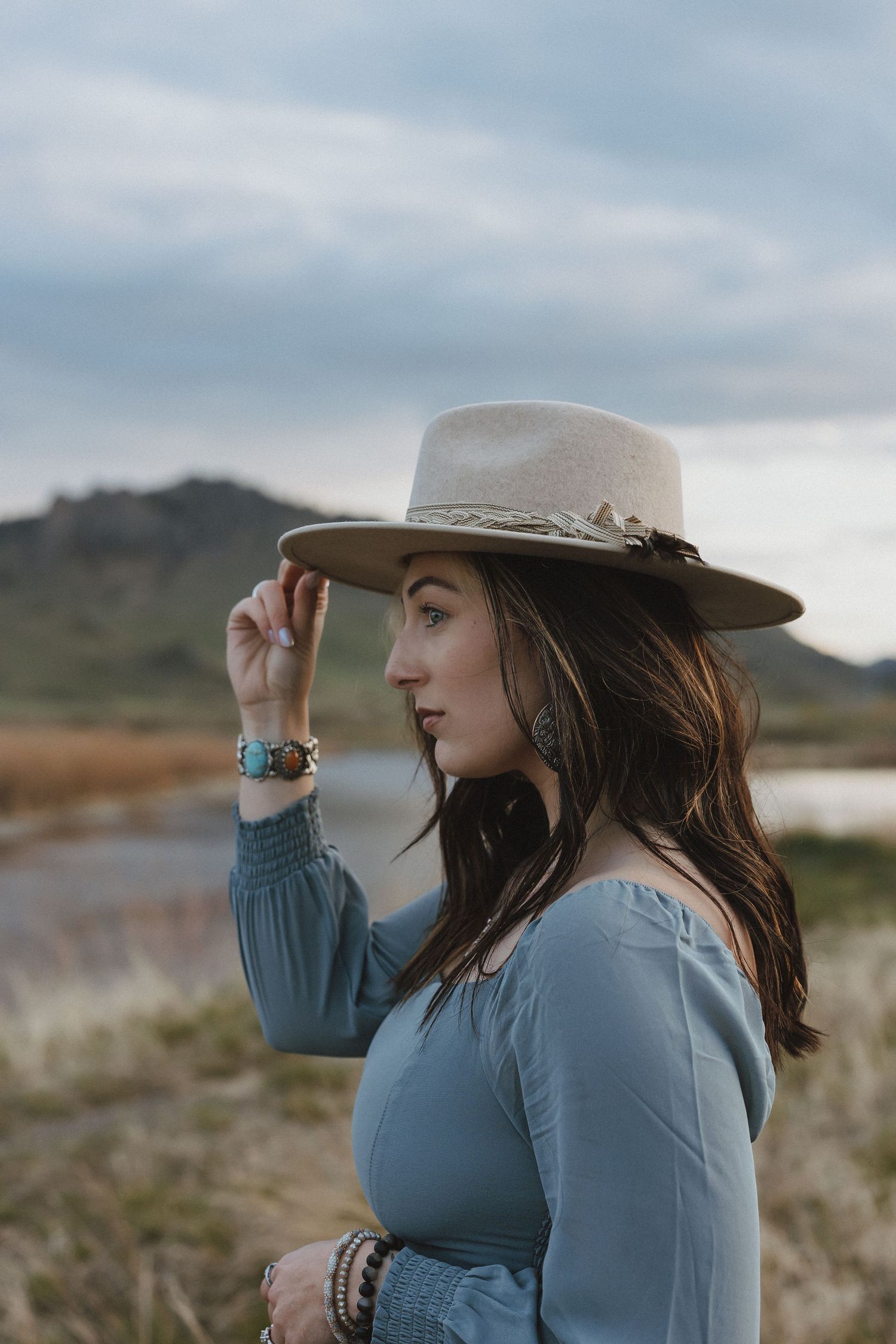 A young woman in a blue dress and wide-brim hat poses outdoors near a lake with mountains in the background.