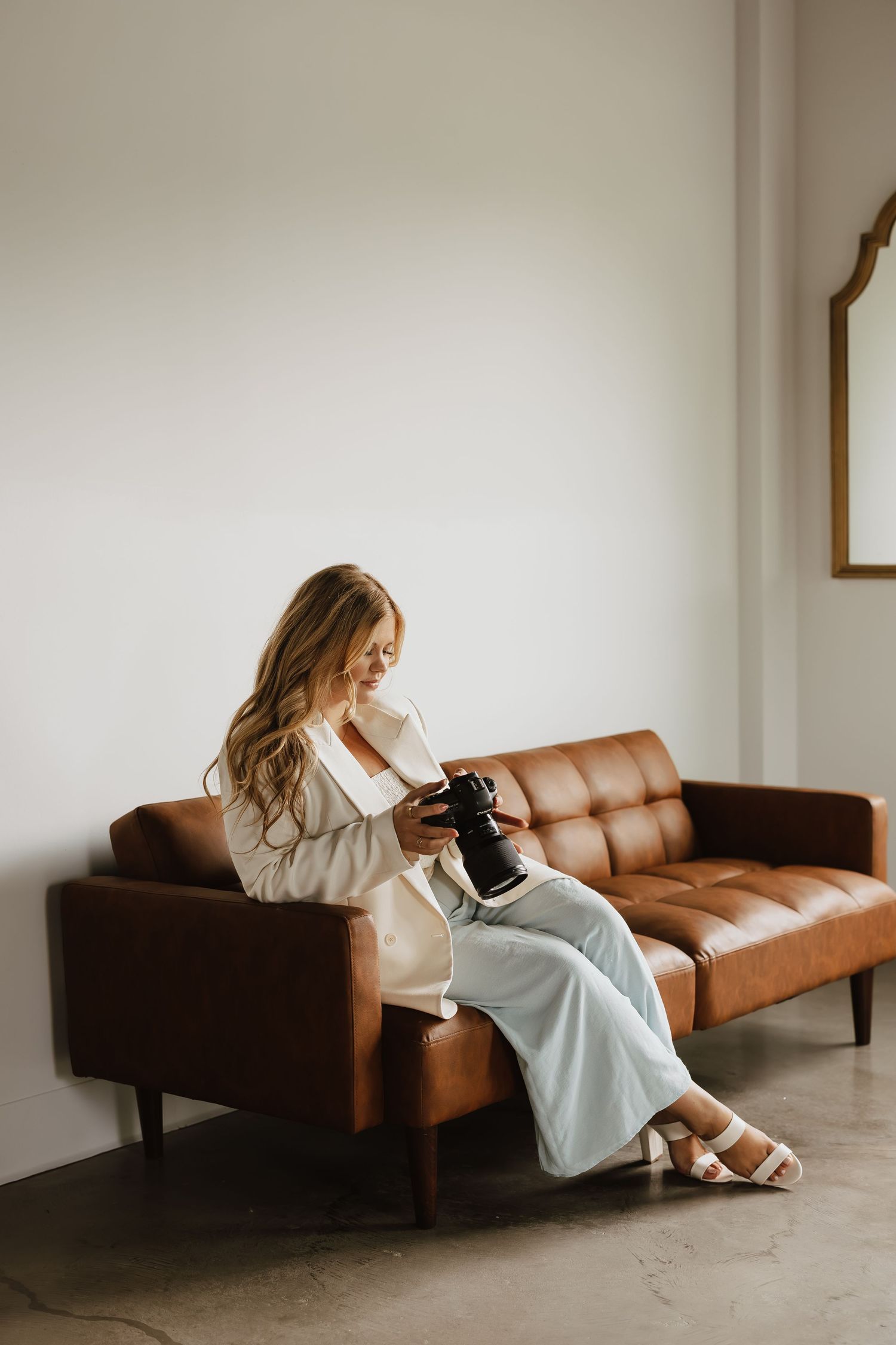 A person in a cozy sweater and wide-leg pants sits on a brown leather sofa while looking at a camera.
