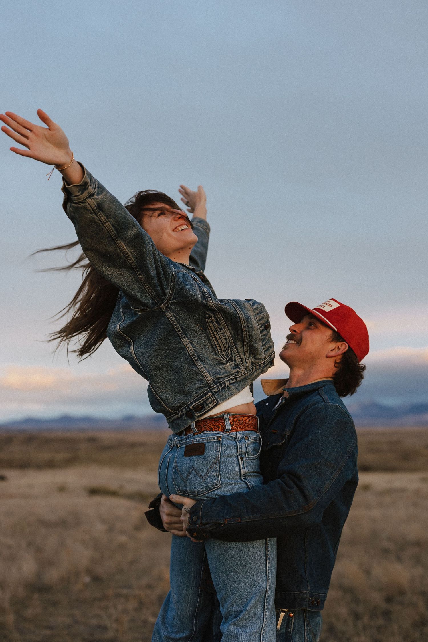 A man in a red cap lifts a woman in a denim jacket outdoors, as she reaches her arms up toward the sky.