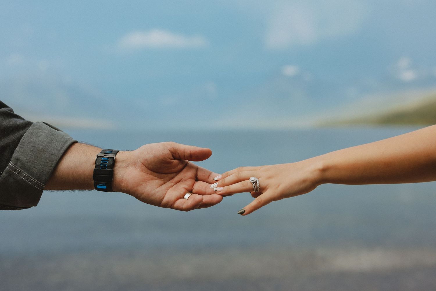 Two hands gently touching, man wearing a watch, woman with rings, against a blurred blue water background.