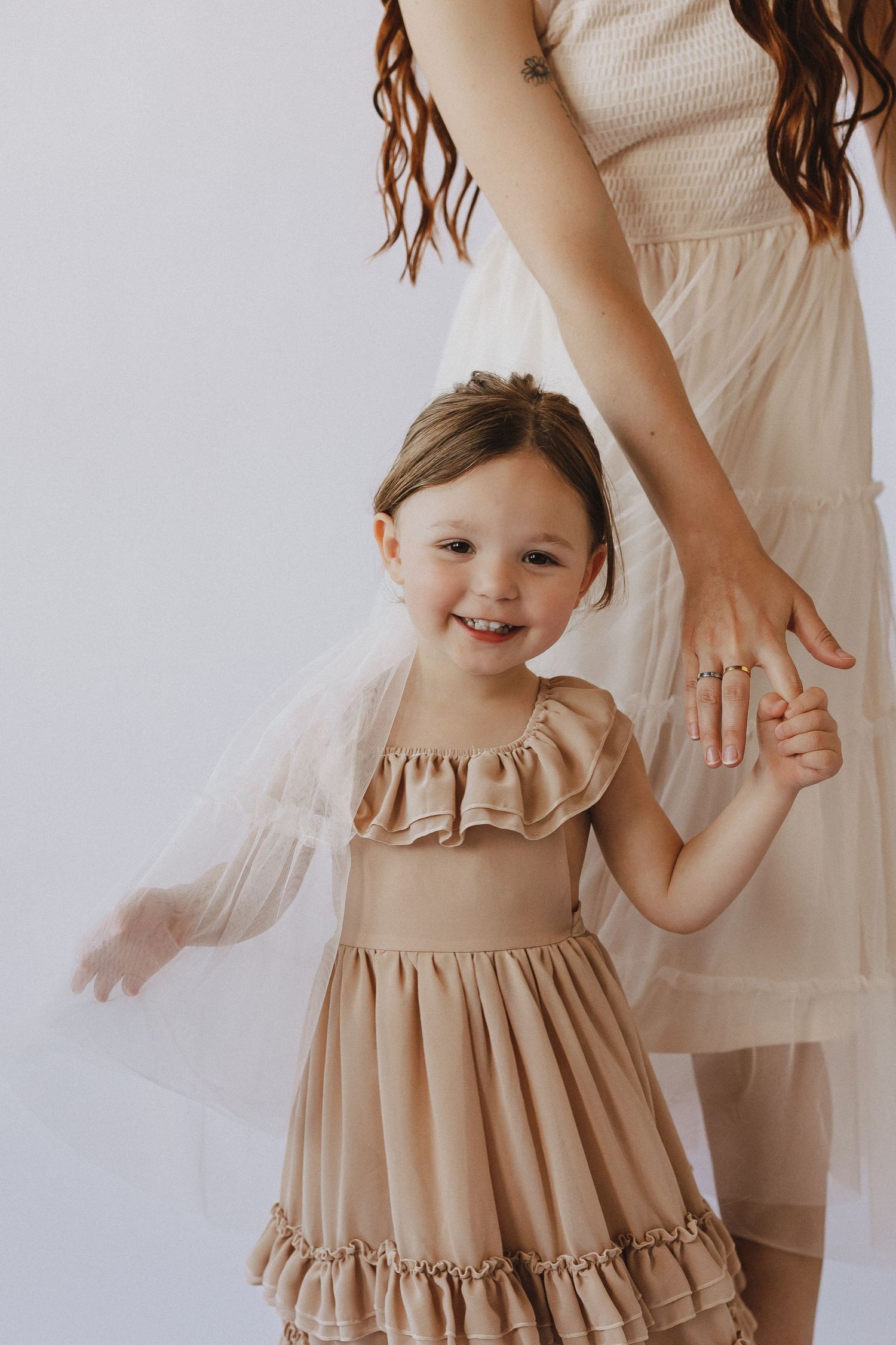 A smiling little girl in a tan ruffle dress holds hands with a woman in a matching beige smocked dress against a white background.