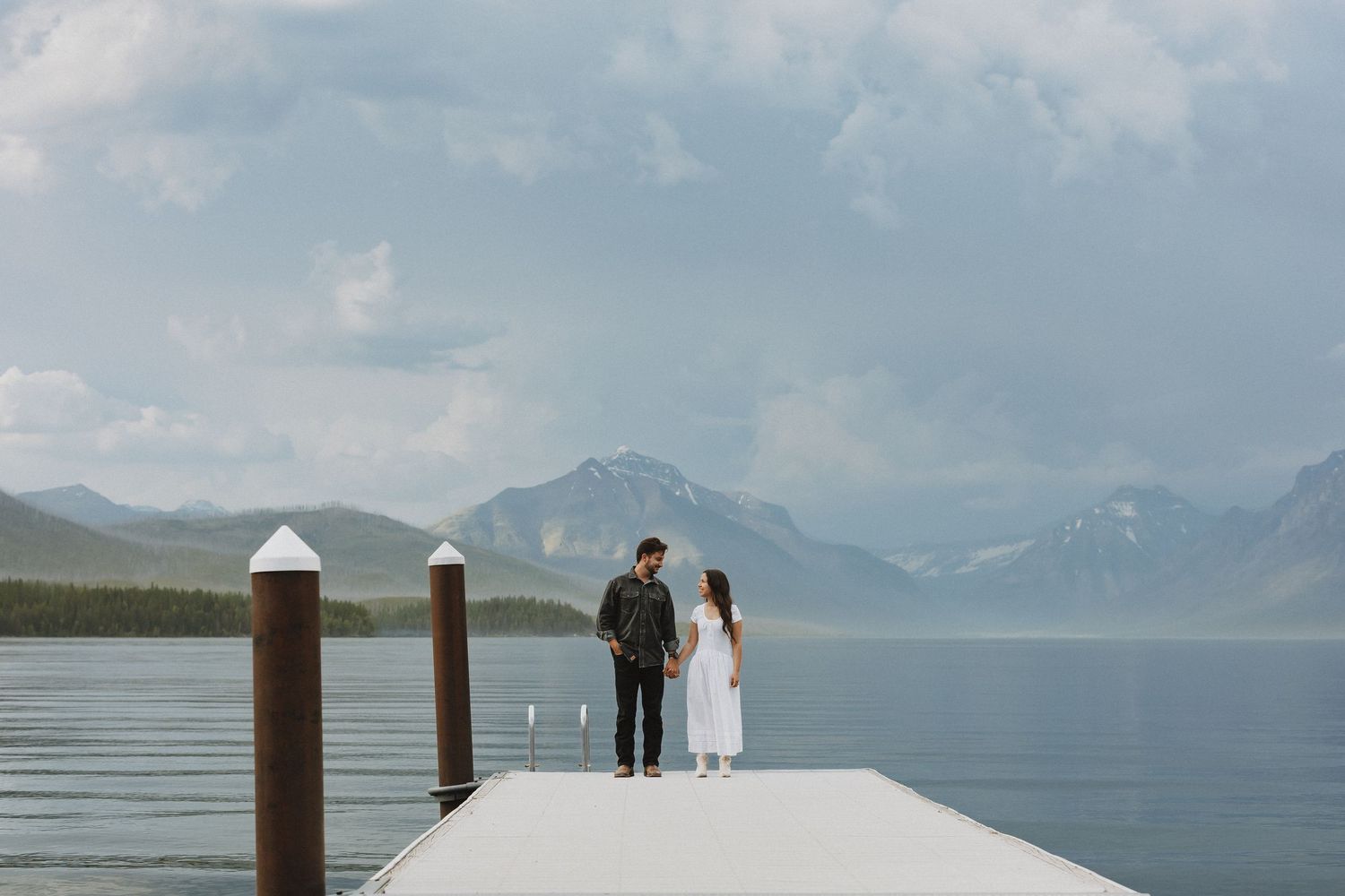 Couple holding hands on a dock at Lake McDonald in Glacier National Park with mountain views.