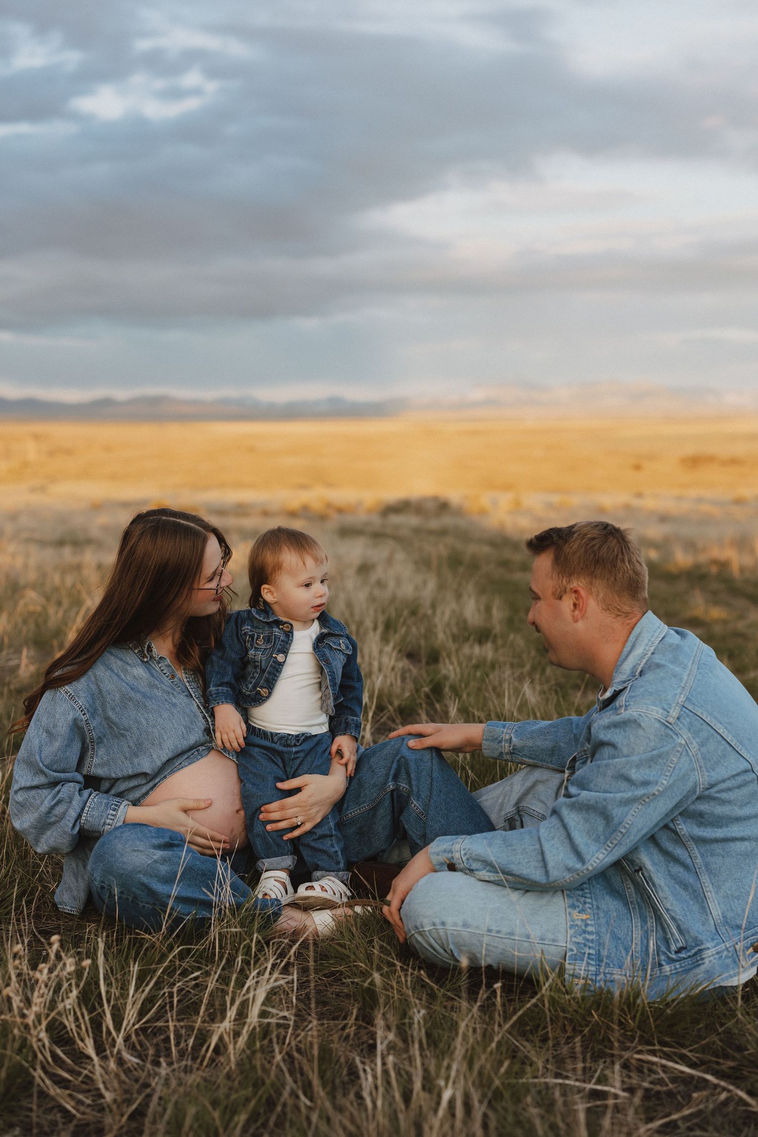 Pregnant woman and man sitting in golden field with toddler, family wearing matching denim outfits at sunset.