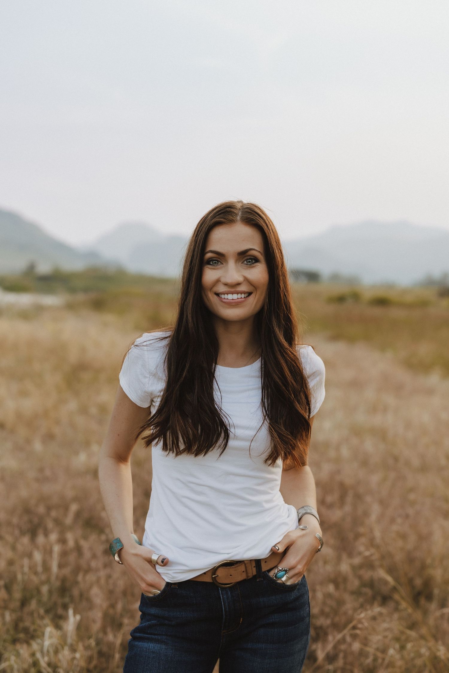 Smiling brunette woman in white t-shirt and jeans standing in a golden field with mountains behind her.