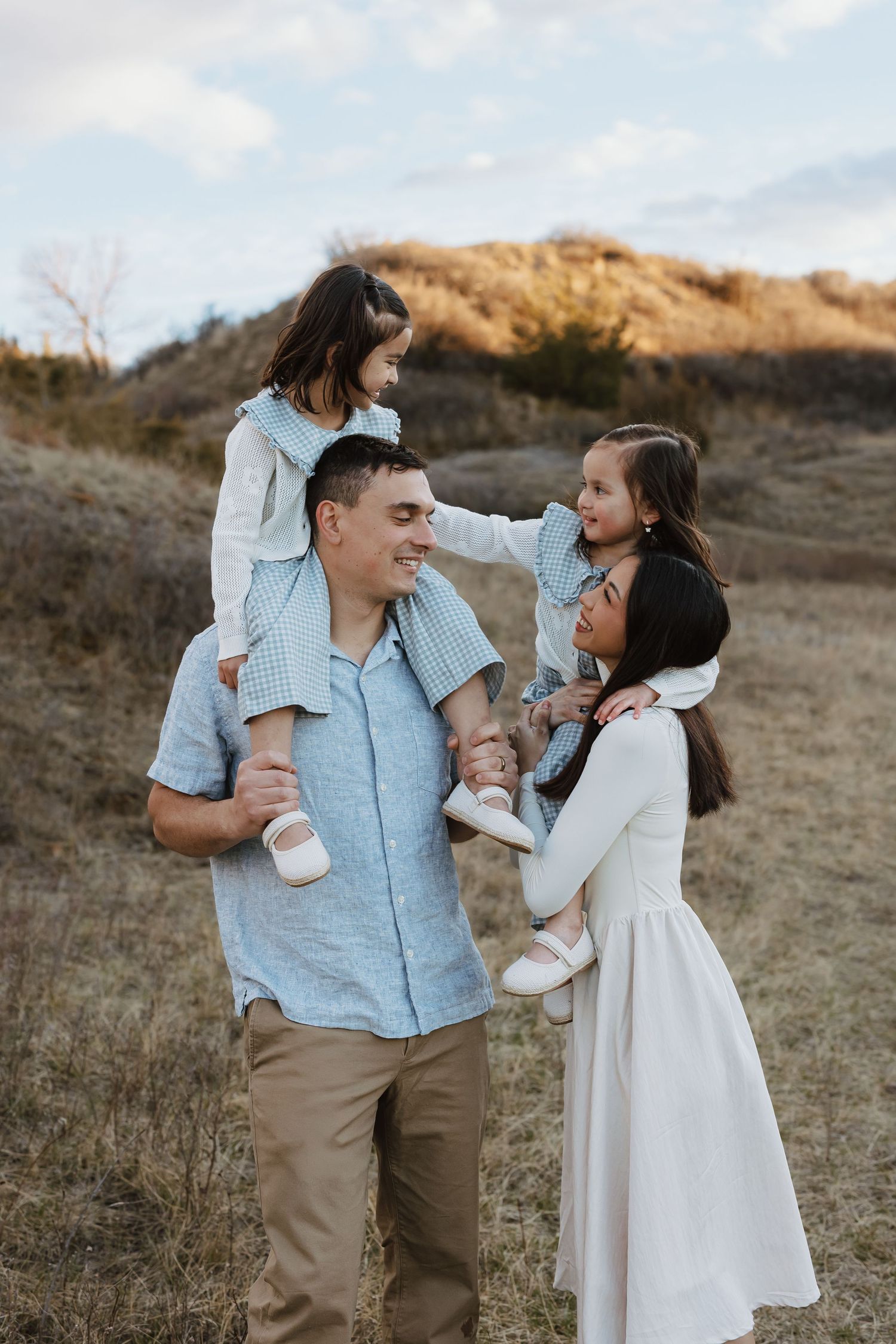 Happy family of four outdoors, dad carrying daughter on shoulders while mom holds other child in golden field.