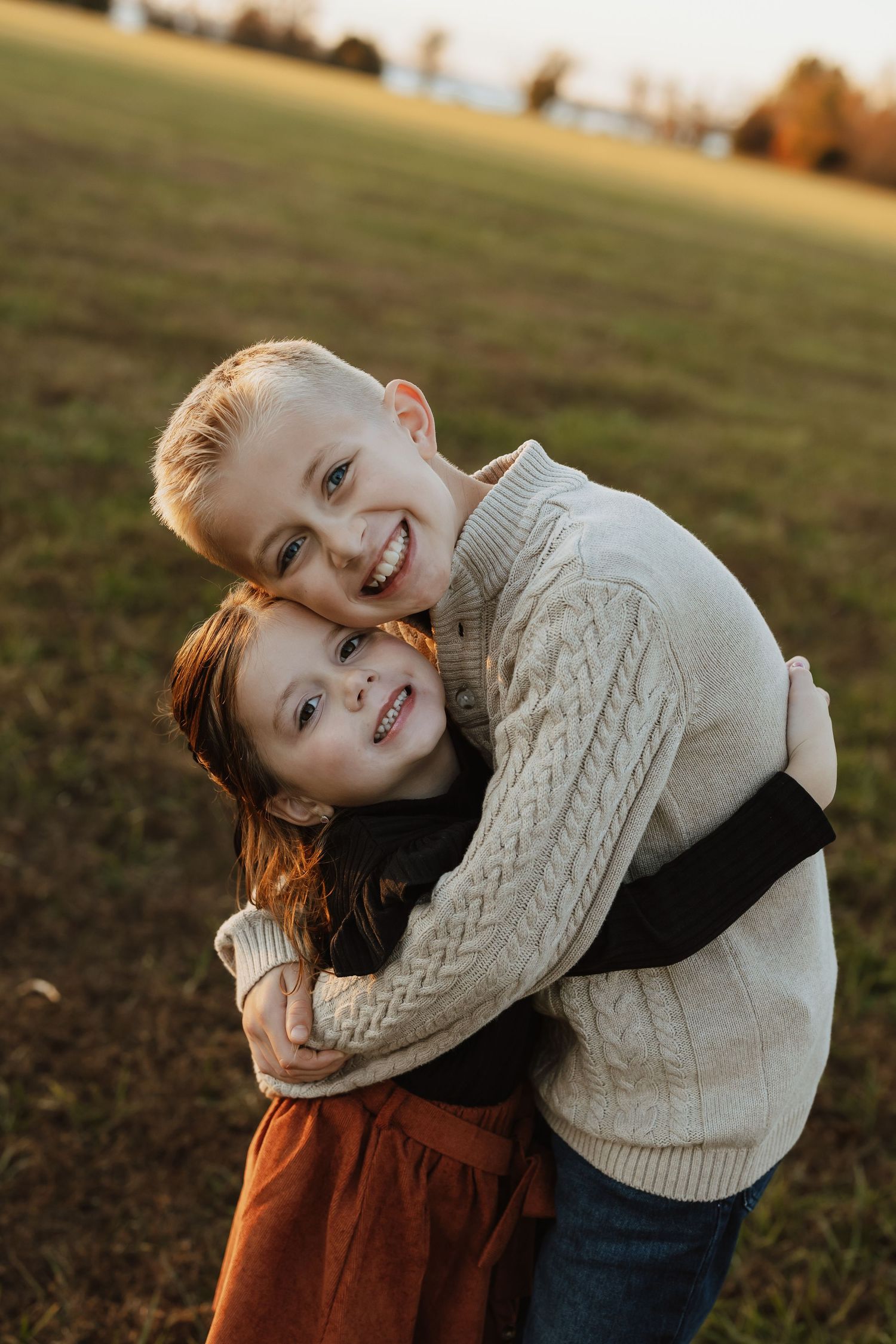 Two people in cozy sweaters embrace playfully in a field at sunset.