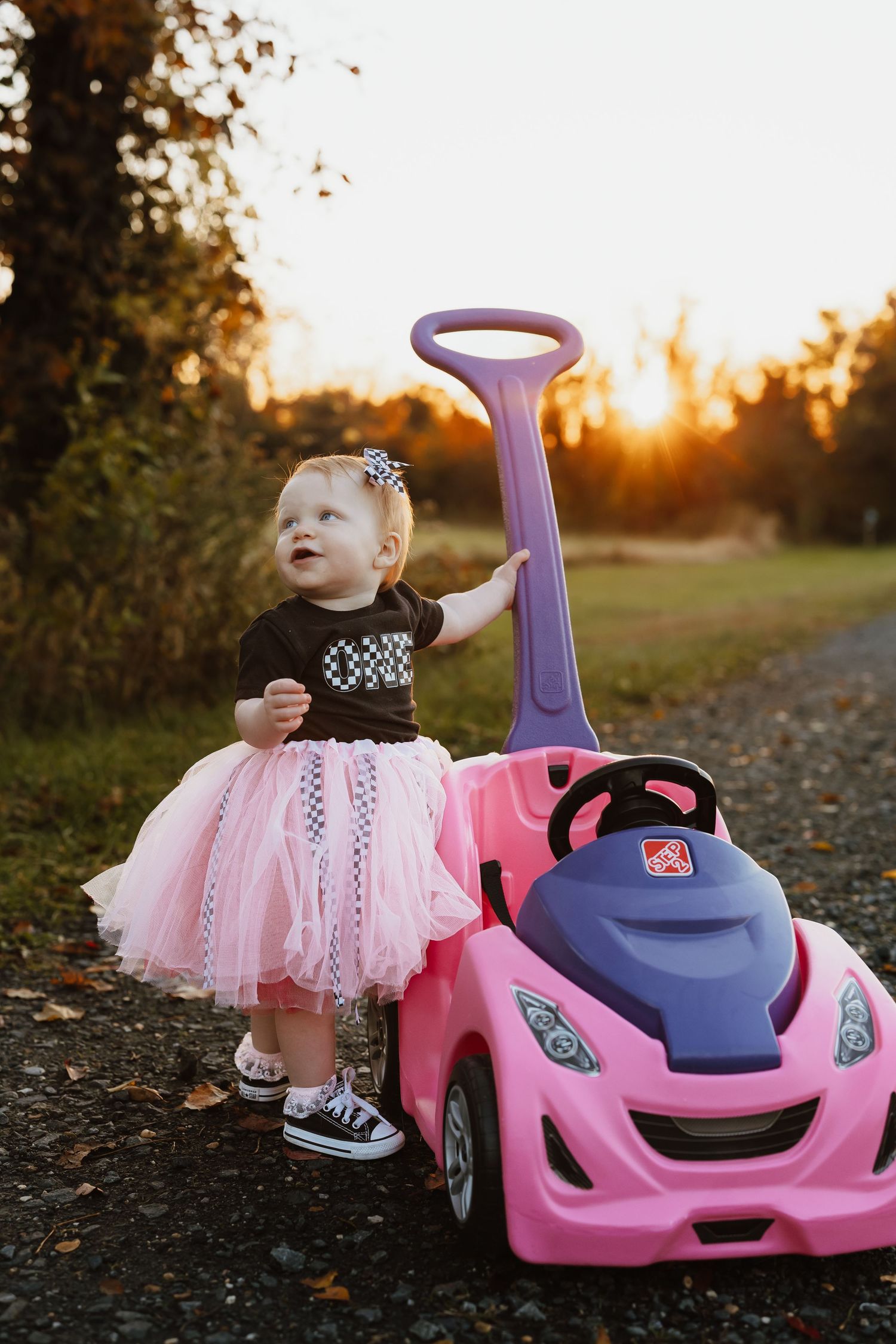A pink toy push car and a small child in a tulle skirt stand on a path during golden hour sunset.
