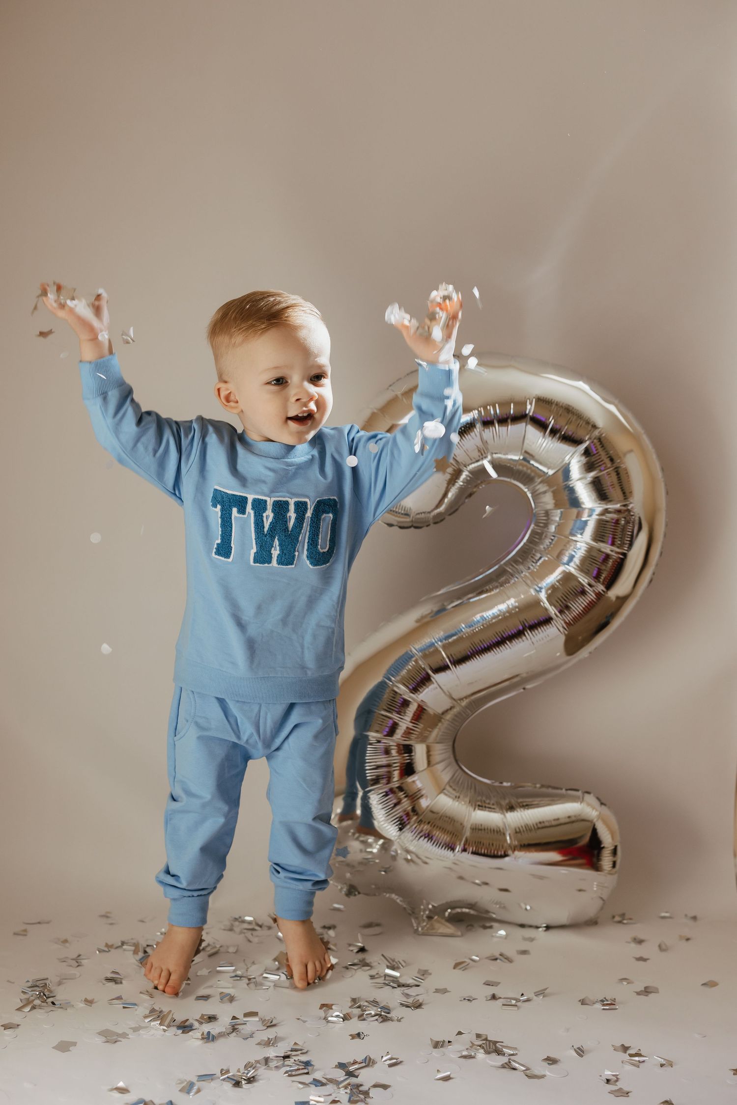 A small child in blue romper plays with a silver number 2 balloon amid scattered confetti on a neutral backdrop.
