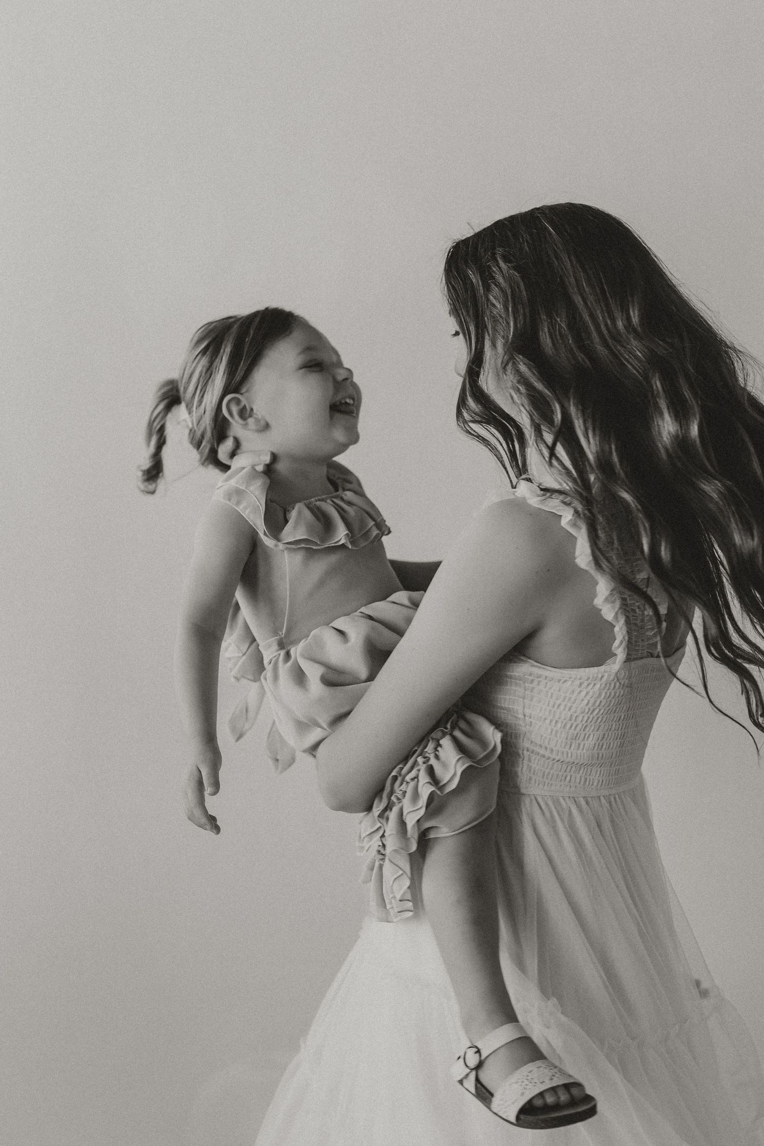 Black and white photo of a woman holding a laughing toddler girl, both wearing dresses, sharing a joyful moment.