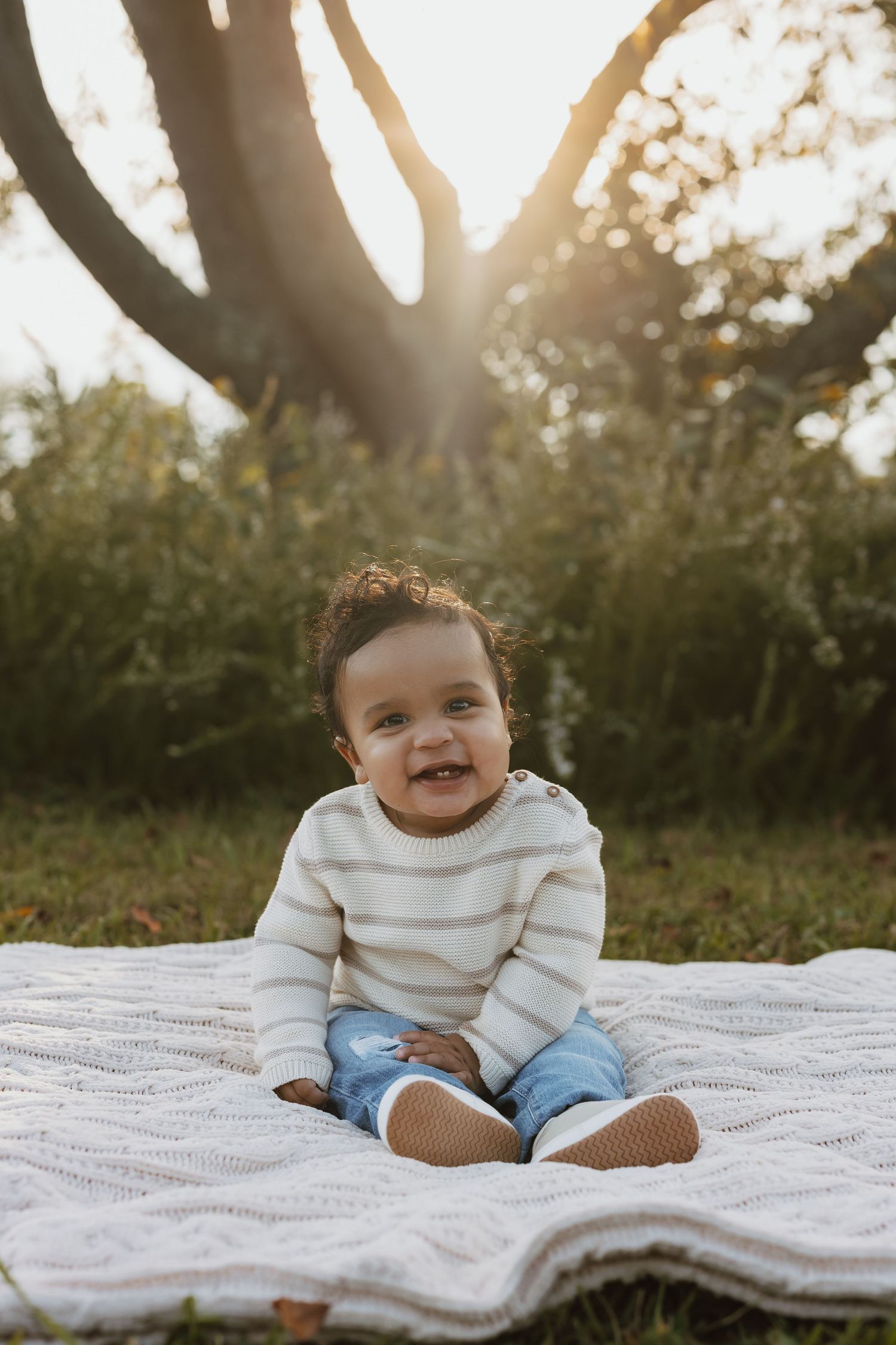 Baby in white sweater and jeans sits on white blanket outdoors during golden hour sunset.