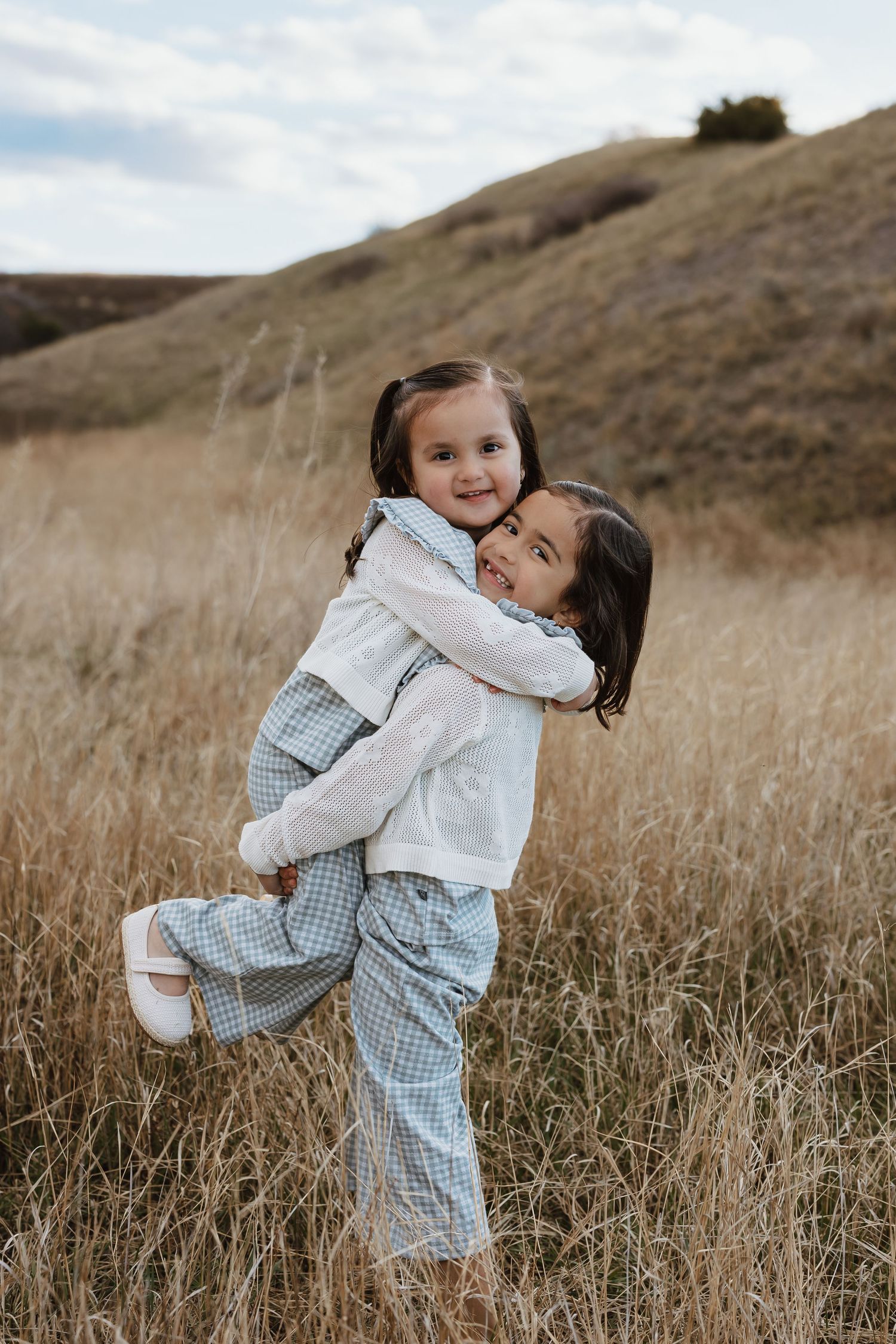 Two young girls hugging in a golden field, one giving the other a piggyback ride, smiling joyfully outdoors.