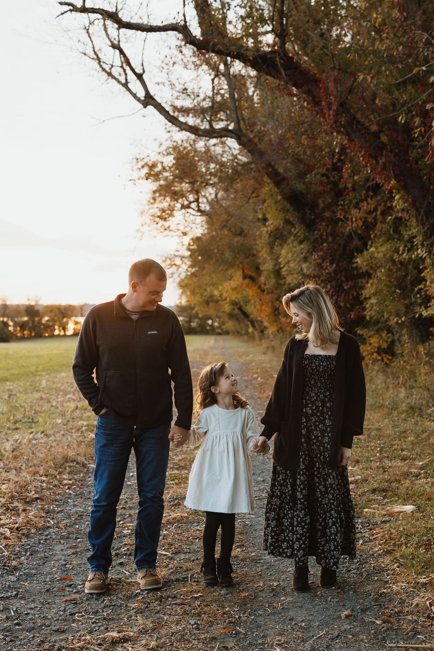 A family of three walks together on a tree-lined path during golden hour sunset.