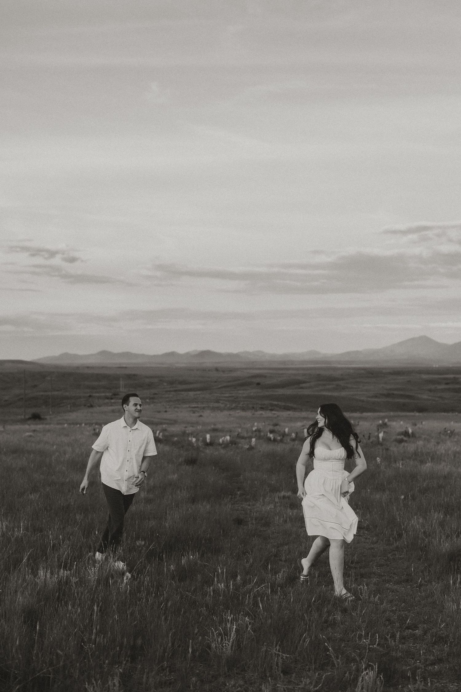Black and white photo of a couple running through a vast open field with mountains in the background.