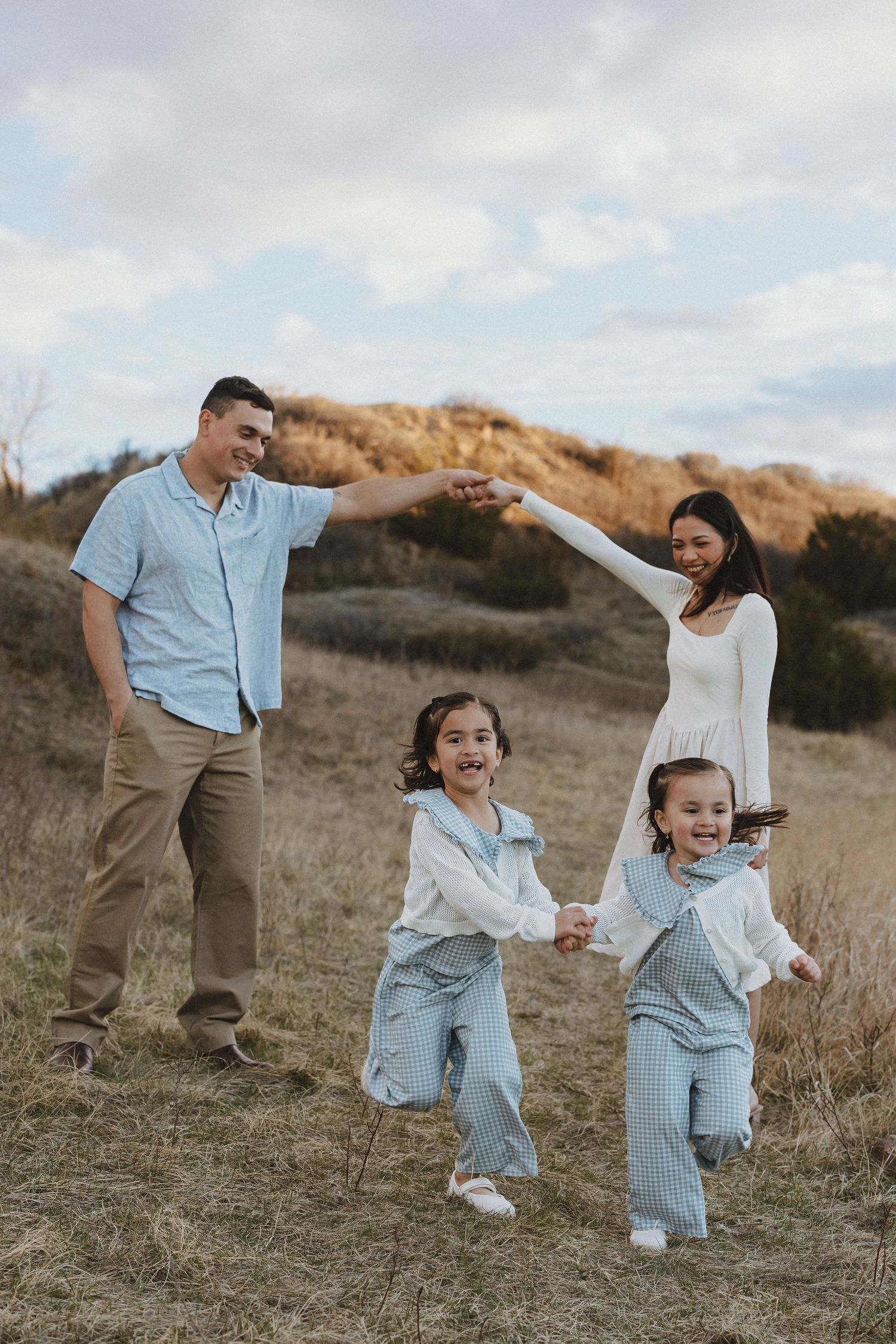 Family of four playing outdoors in a golden field, parents forming an arch while two young girls in matching outfits run and laugh.