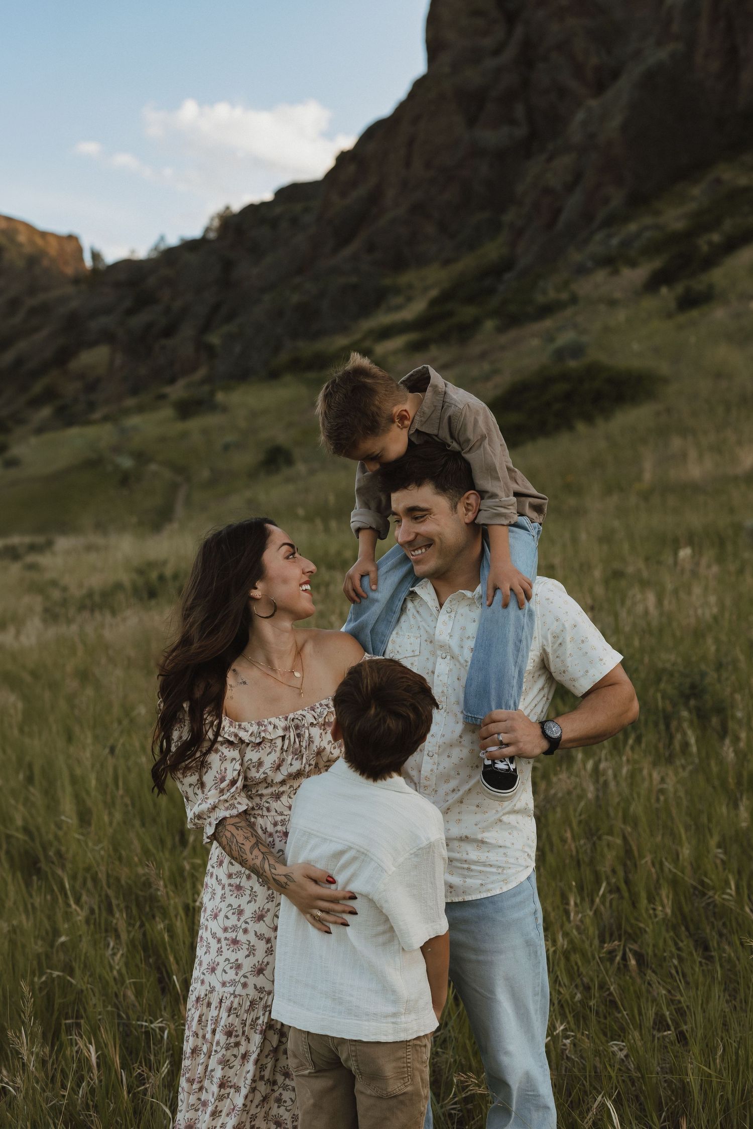 A joyful family of four laughs together in a lush green meadow with rocky mountains in the background.
