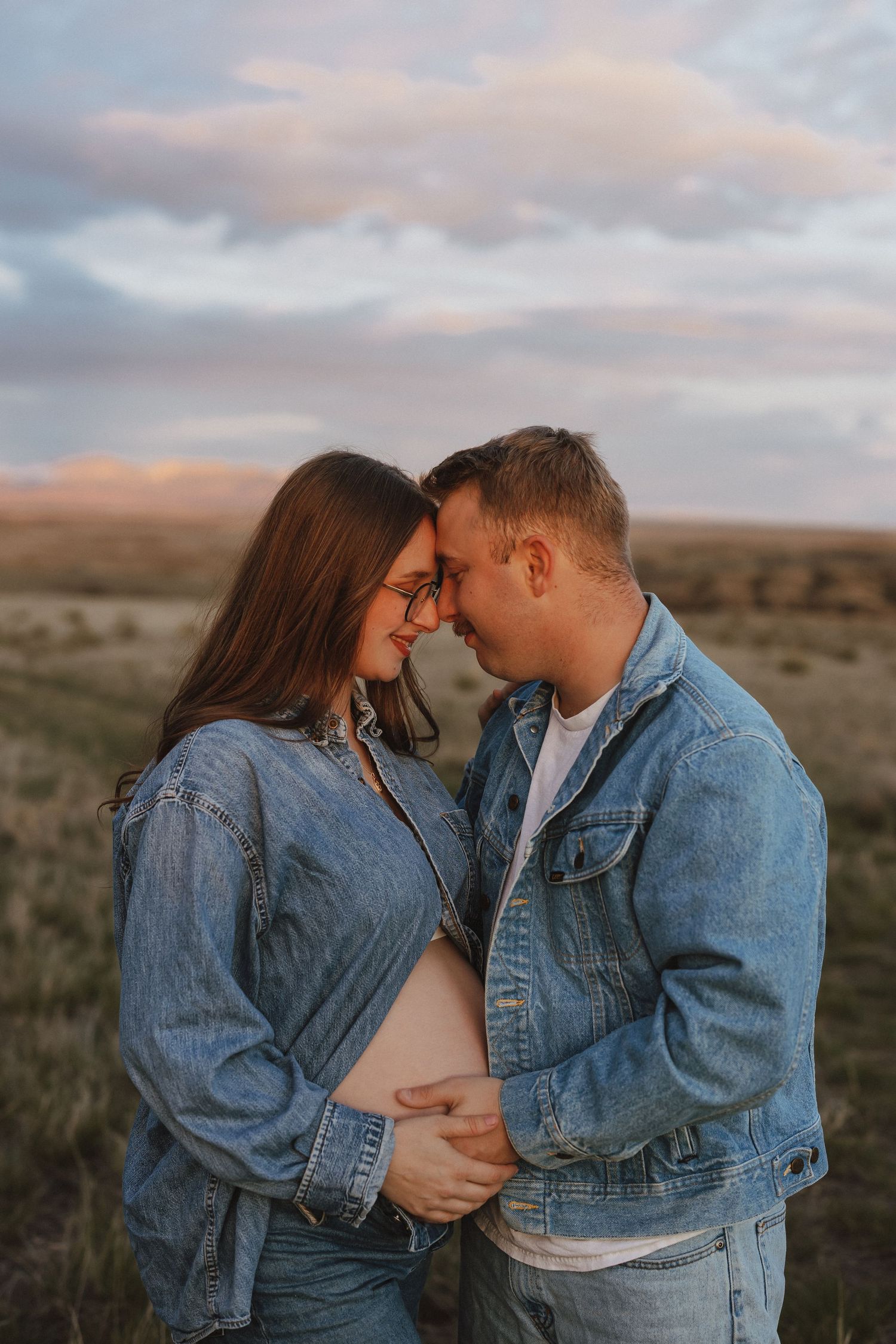 Couple in matching denim jackets share a tender moment outdoors, cradling a baby bump at sunset.