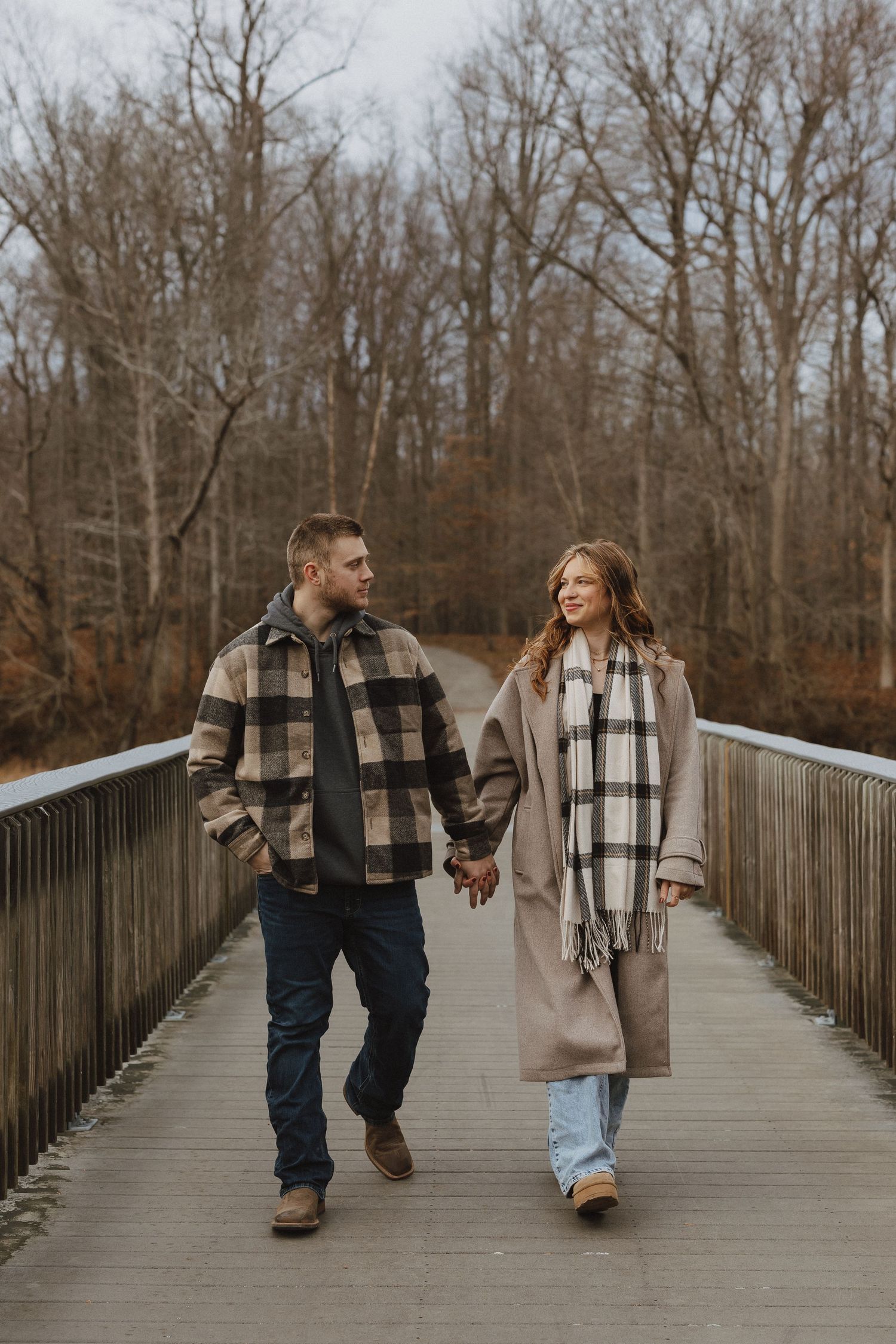 Couple holding hands walking on a wooden bridge in autumn, wearing plaid coats and scarves among bare trees.