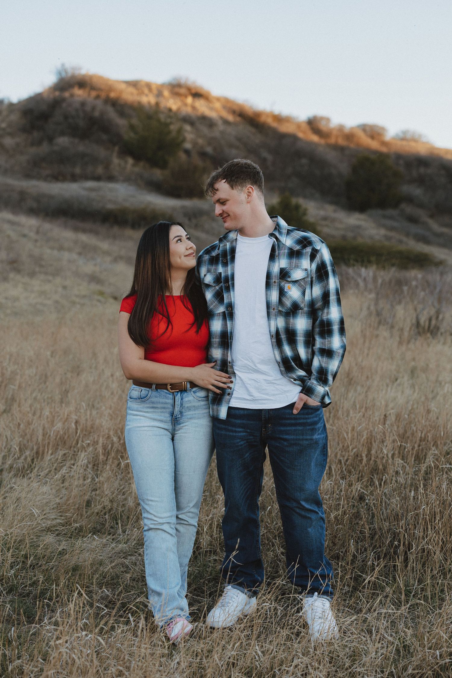 A smiling couple stands in a golden field, woman in red top and jeans, man in plaid shirt, with hills behind.