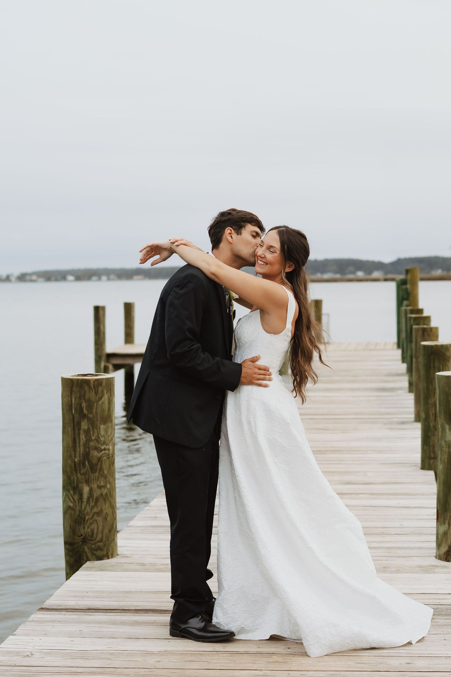 Bride and groom share a romantic kiss on a wooden dock overlooking calm water on their wedding day.