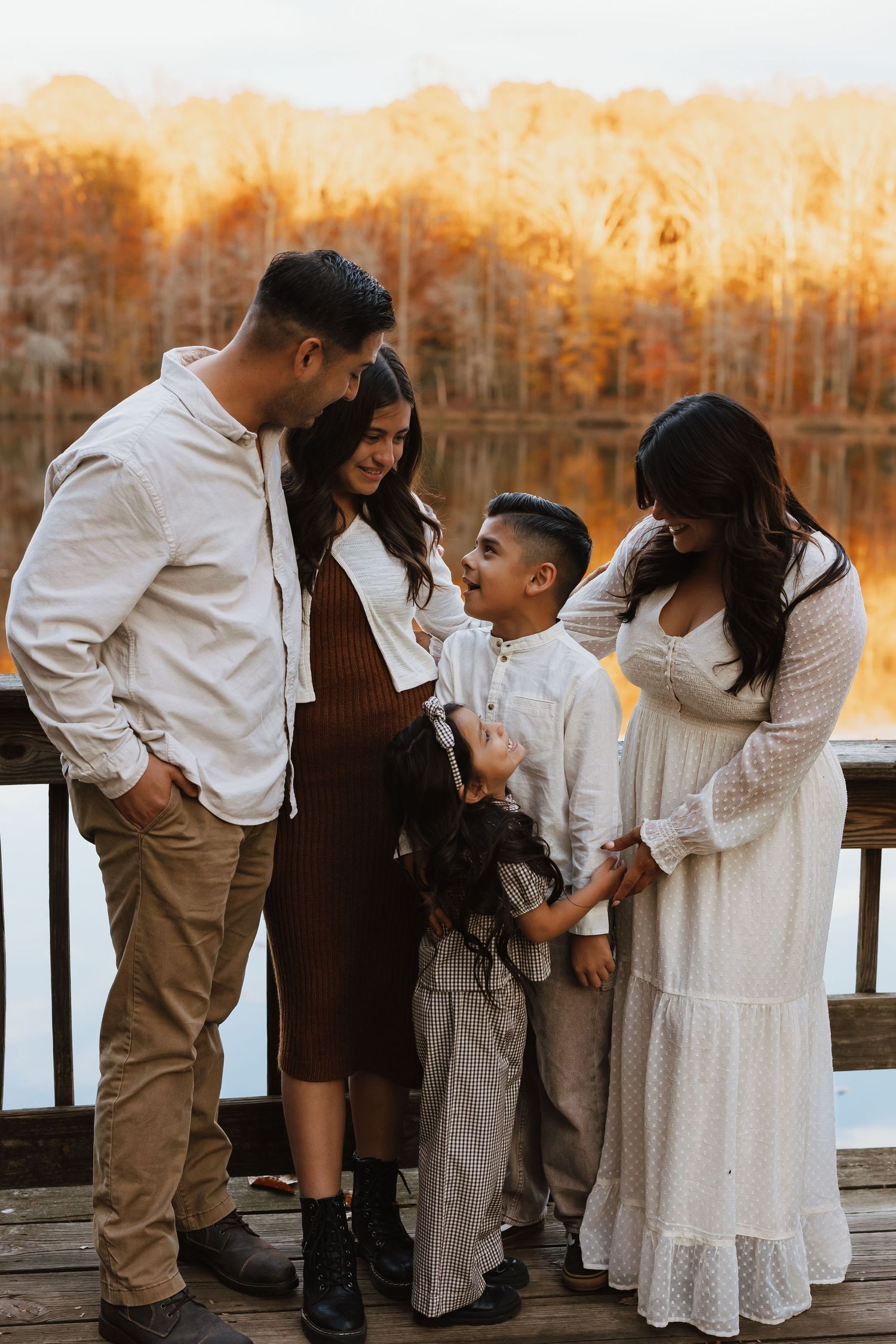 Family dressed in white and neutral tones share a tender moment on wooden dock during golden autumn sunset.