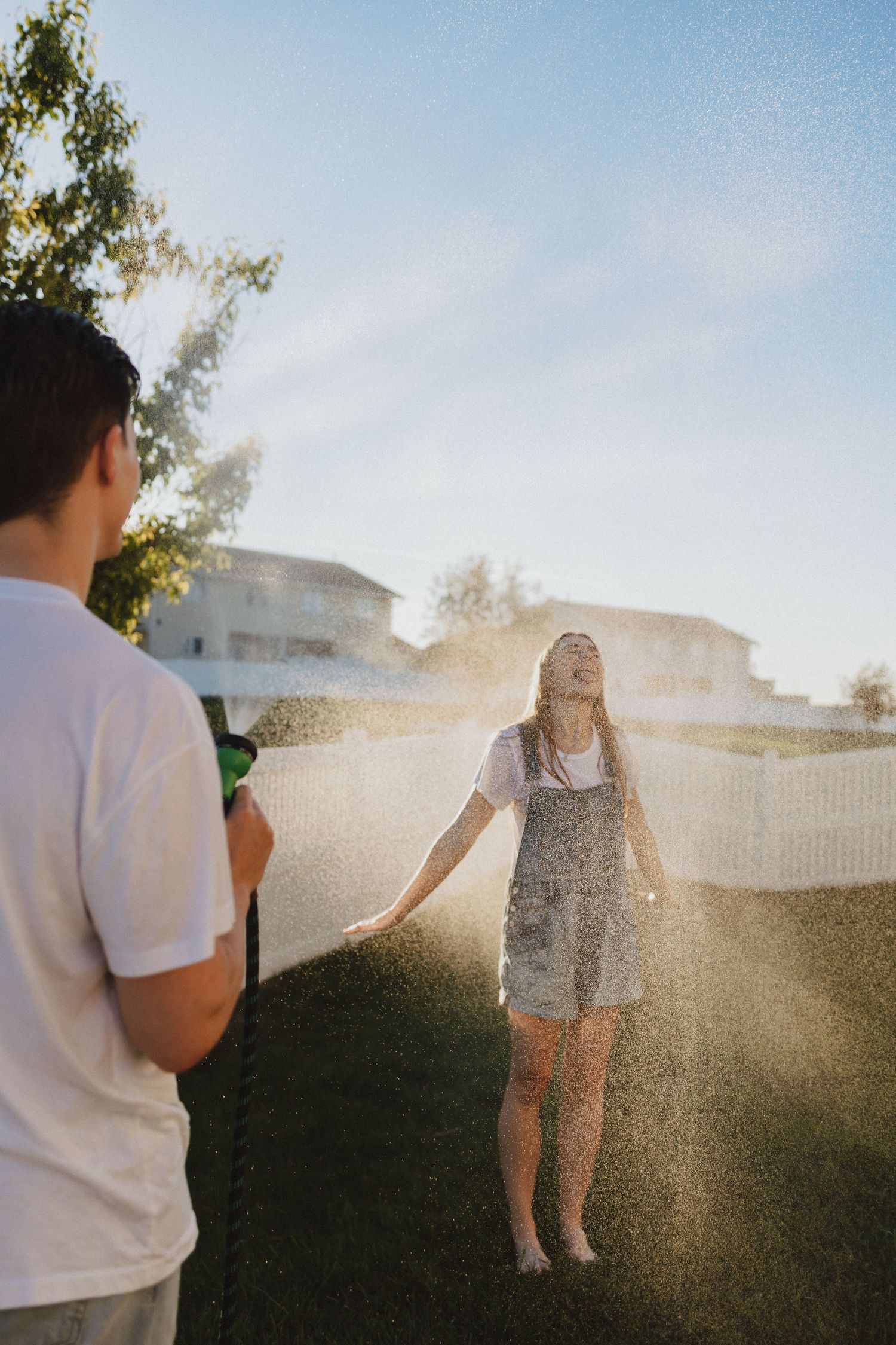 A boy sprays a laughing girl with a garden hose in a sunny backyard, both wearing casual summer clothes.