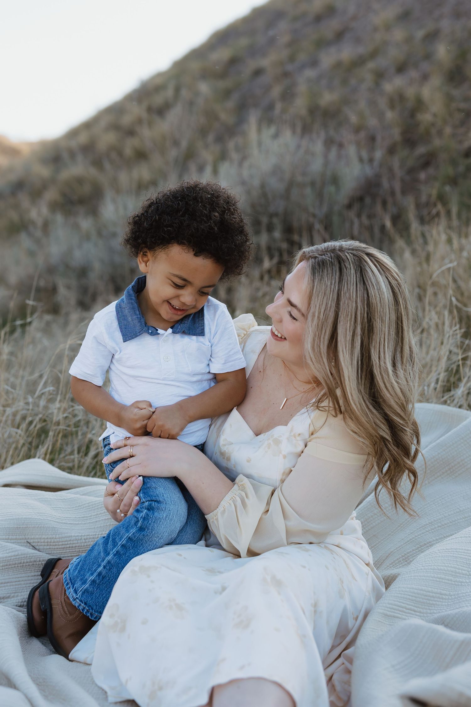 A smiling blonde woman in a floral dress sits on a blanket outdoors with a happy curly-haired boy in denim.