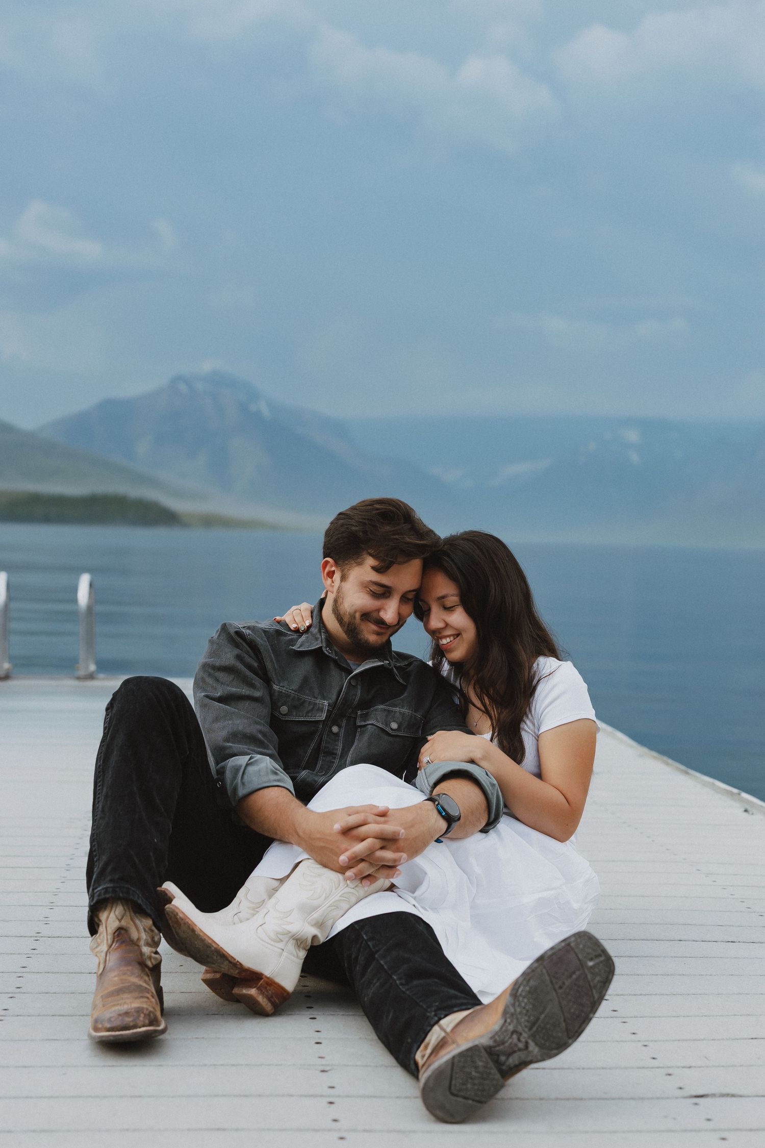 Couple sitting on a dock embracing, with mountains and a lake in the background on a cloudy day.