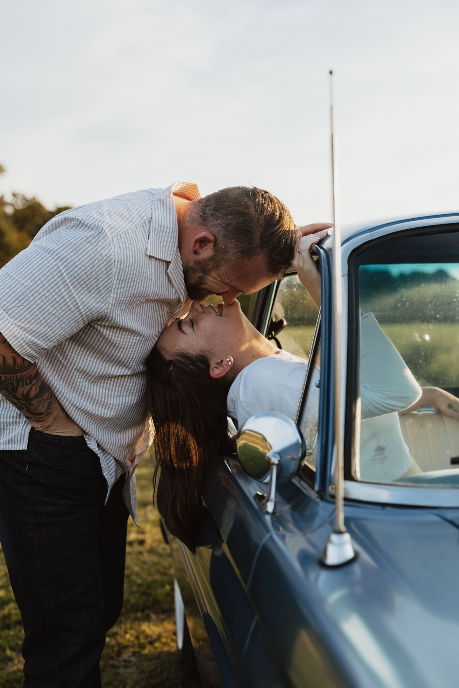A romantic couple shares a tender moment leaning against a vintage car in warm sunlight.